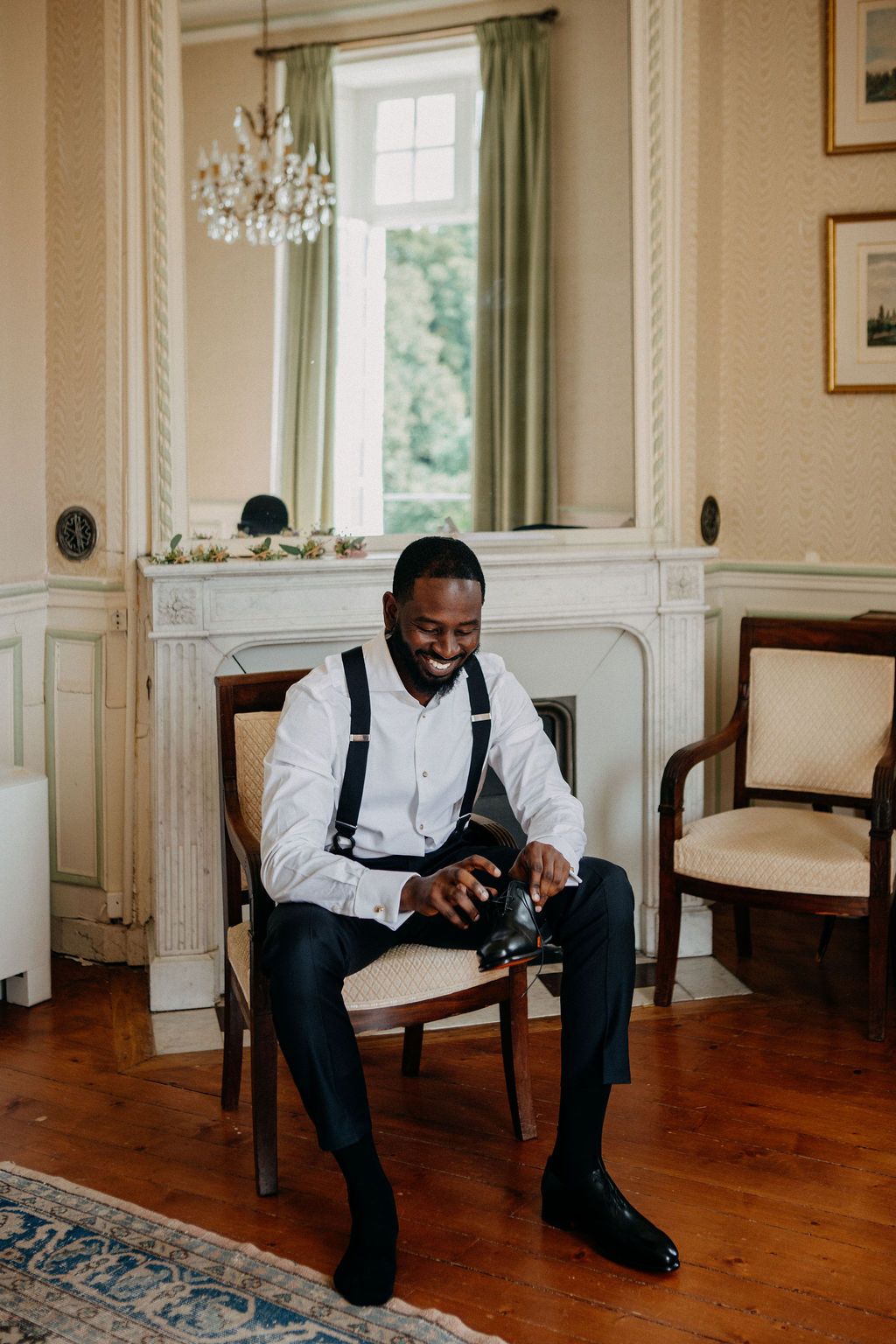 A dapper groom in white shirt and suspenders sits in an elegant chair putting on his dress shoes.