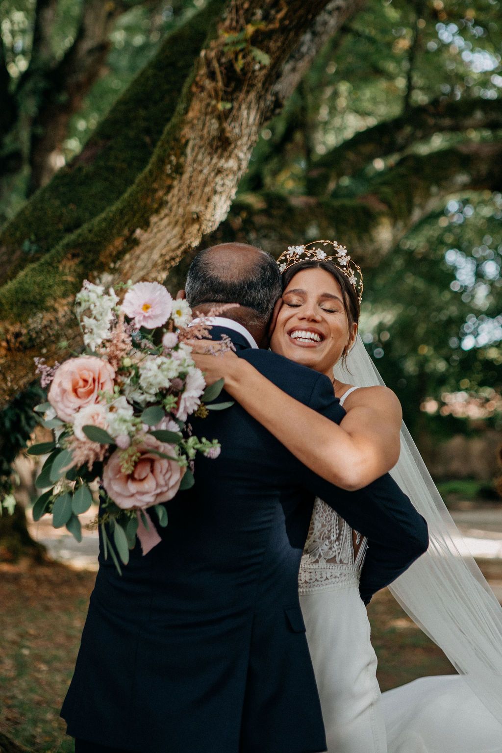 Smiling bride in white gown and floral crown embraces a man while holding a pink bouquet under a mossy tree.