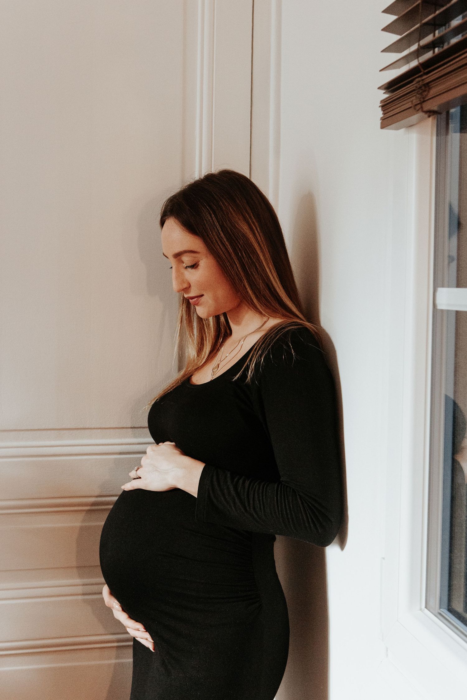 Pregnant woman in black dress gently holding baby bump while standing by window.