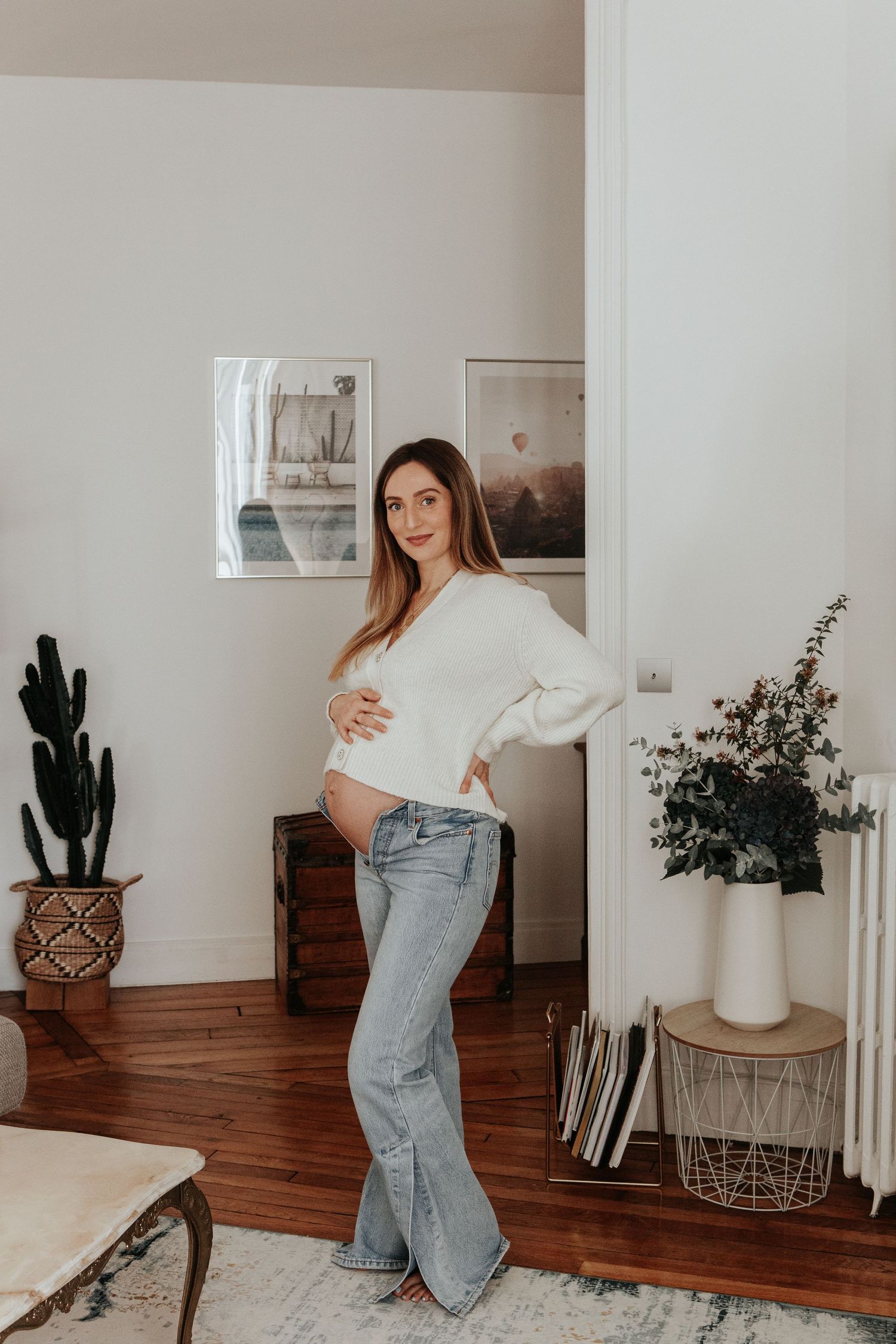 Smiling pregnant woman in white sweater and jeans holding baby bump in bright living room.