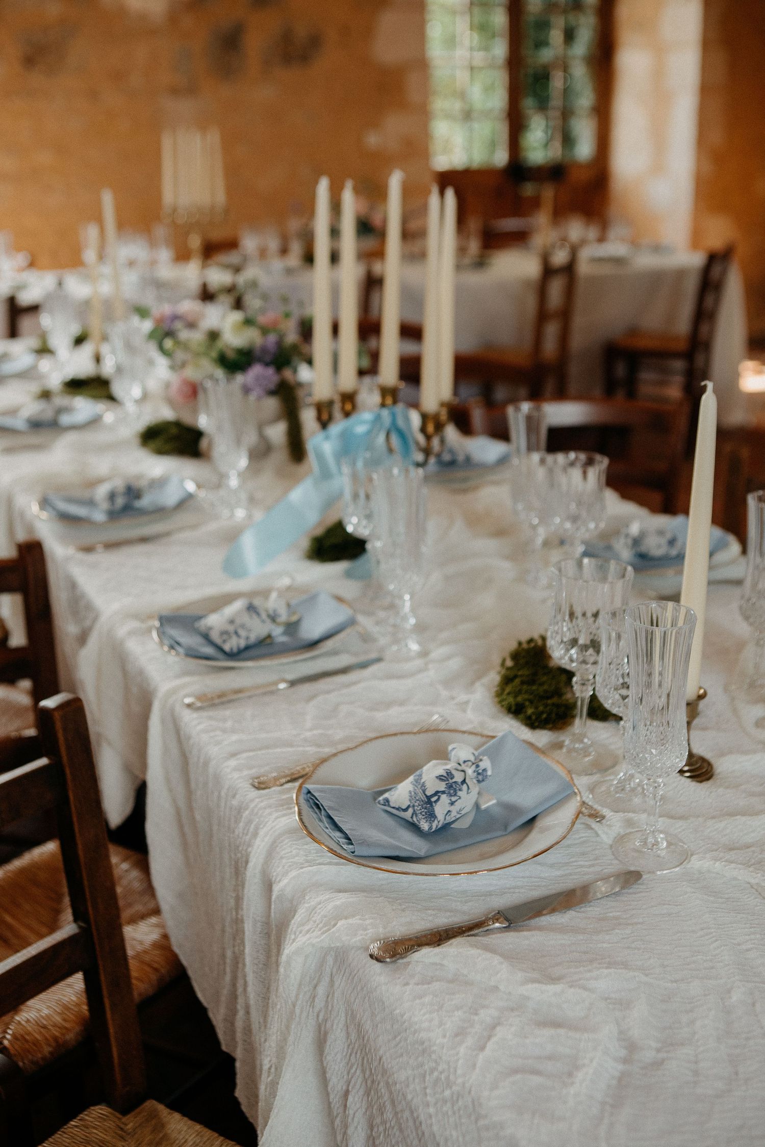 Elegant wedding table setting with blue napkins, crystal glasses, white candles, and floral centerpieces on a white lace tablecloth.
