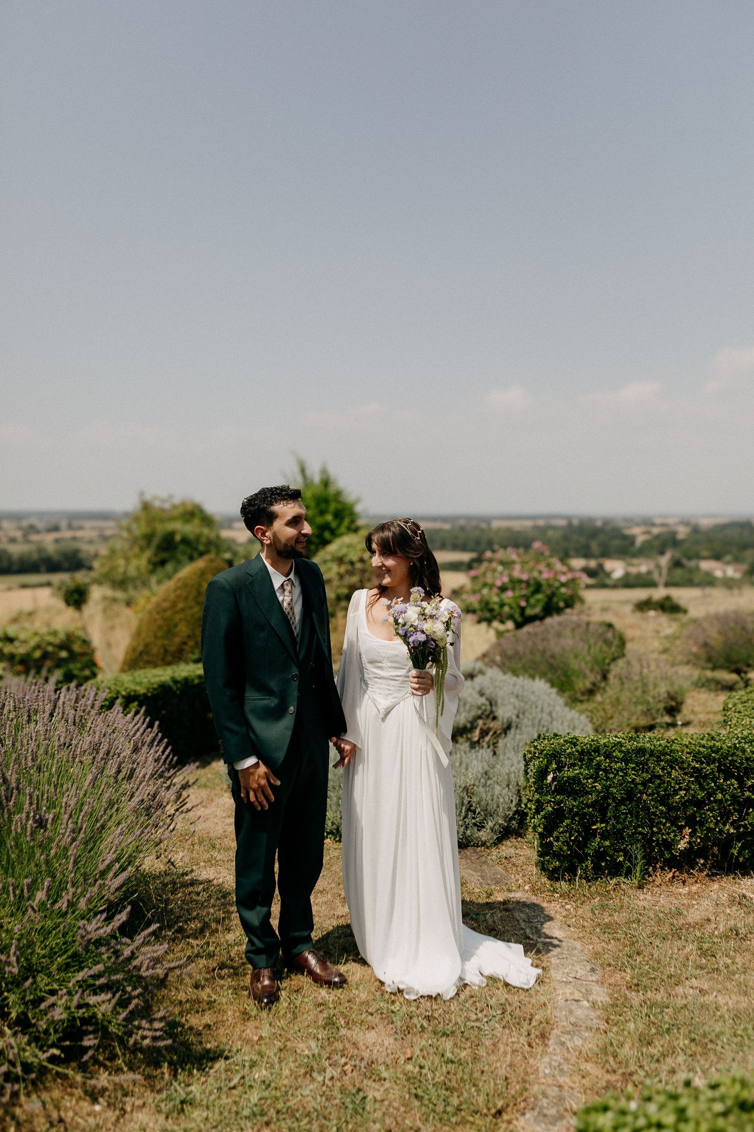 Bride and groom stand together in a sunny garden with lavender and countryside views, bride holding a bouquet.