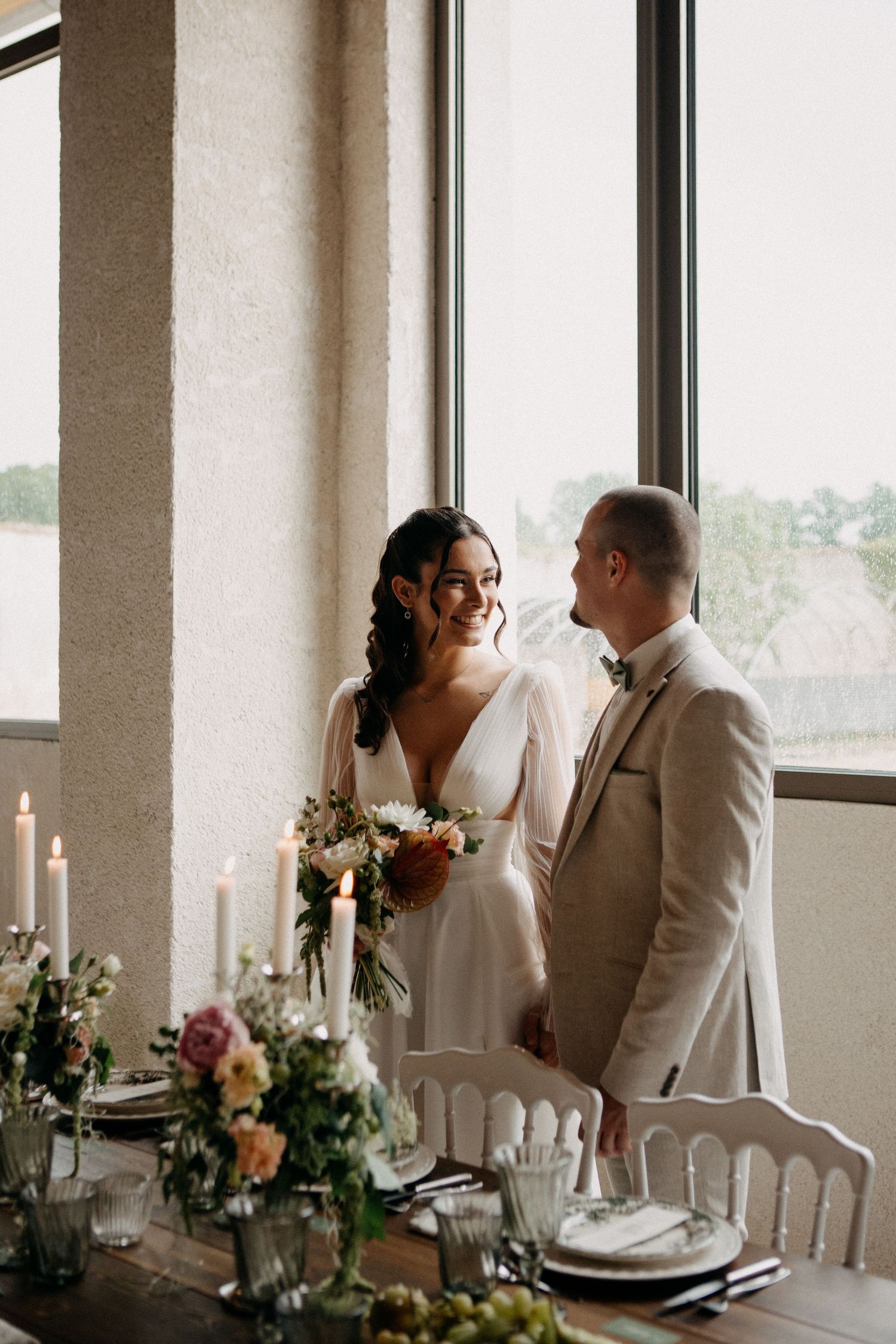 Smiling bride in white gown and groom in beige suit standing by candlelit wedding reception table.