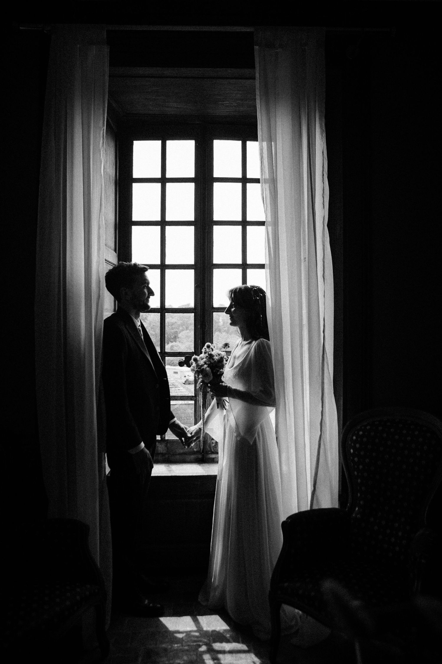 Black and white photo of bride and groom holding hands by a large window with white curtains on their wedding day.