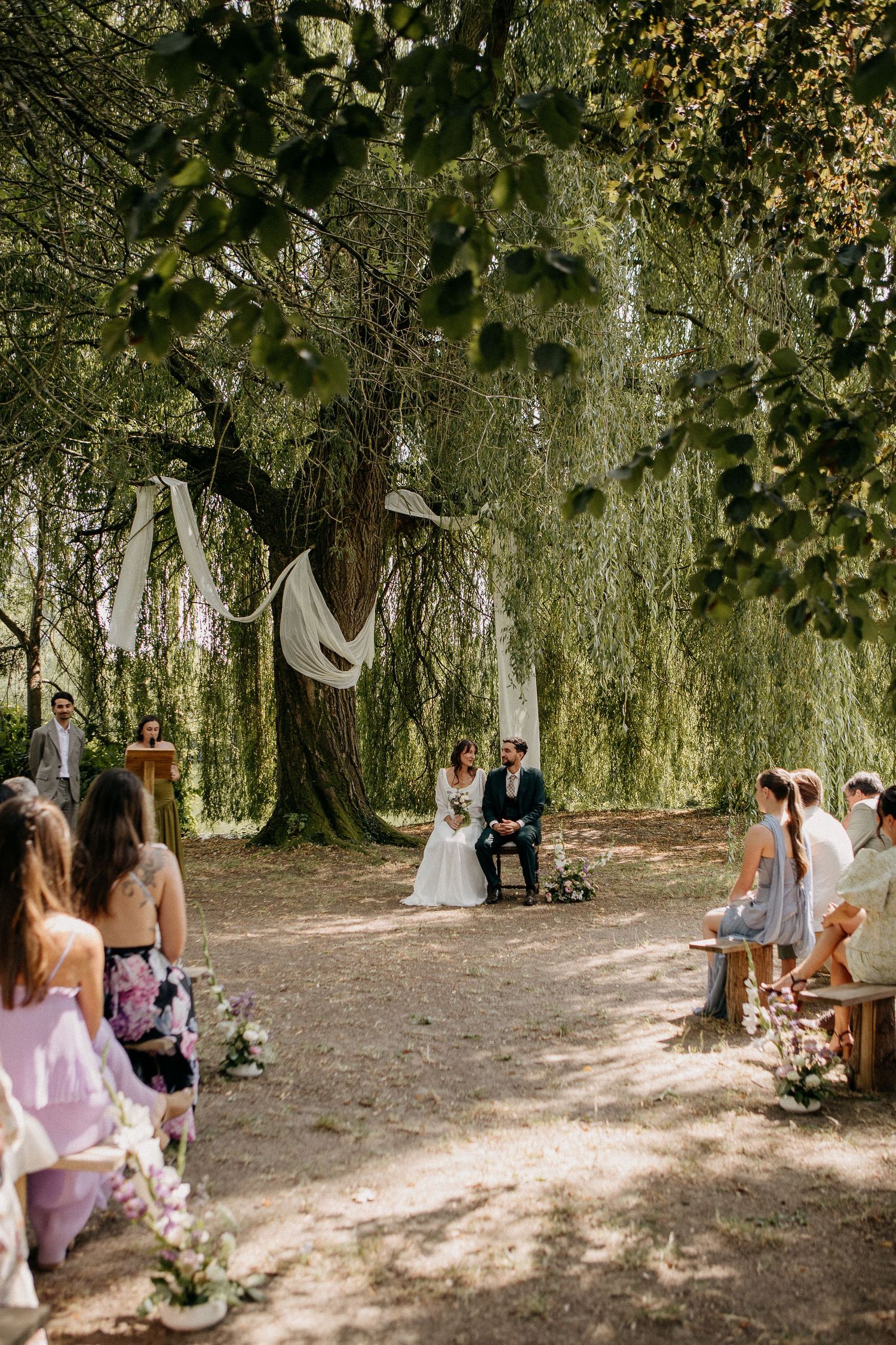 Outdoor wedding ceremony under large willow trees with couple seated at altar and guests on wooden benches in dappled sunlight.