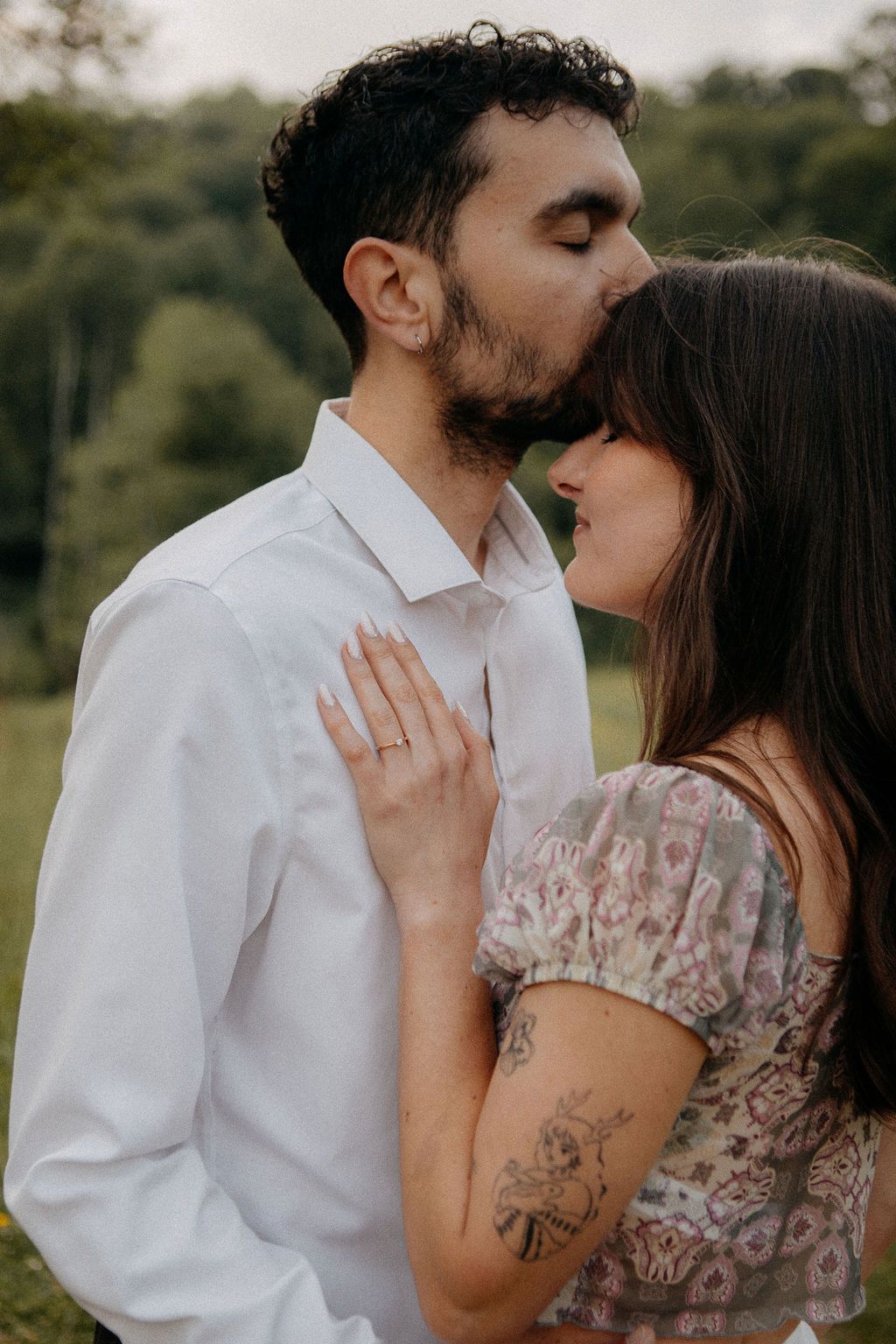 A bearded man tenderly kisses a smiling woman on the forehead in a romantic outdoor portrait.