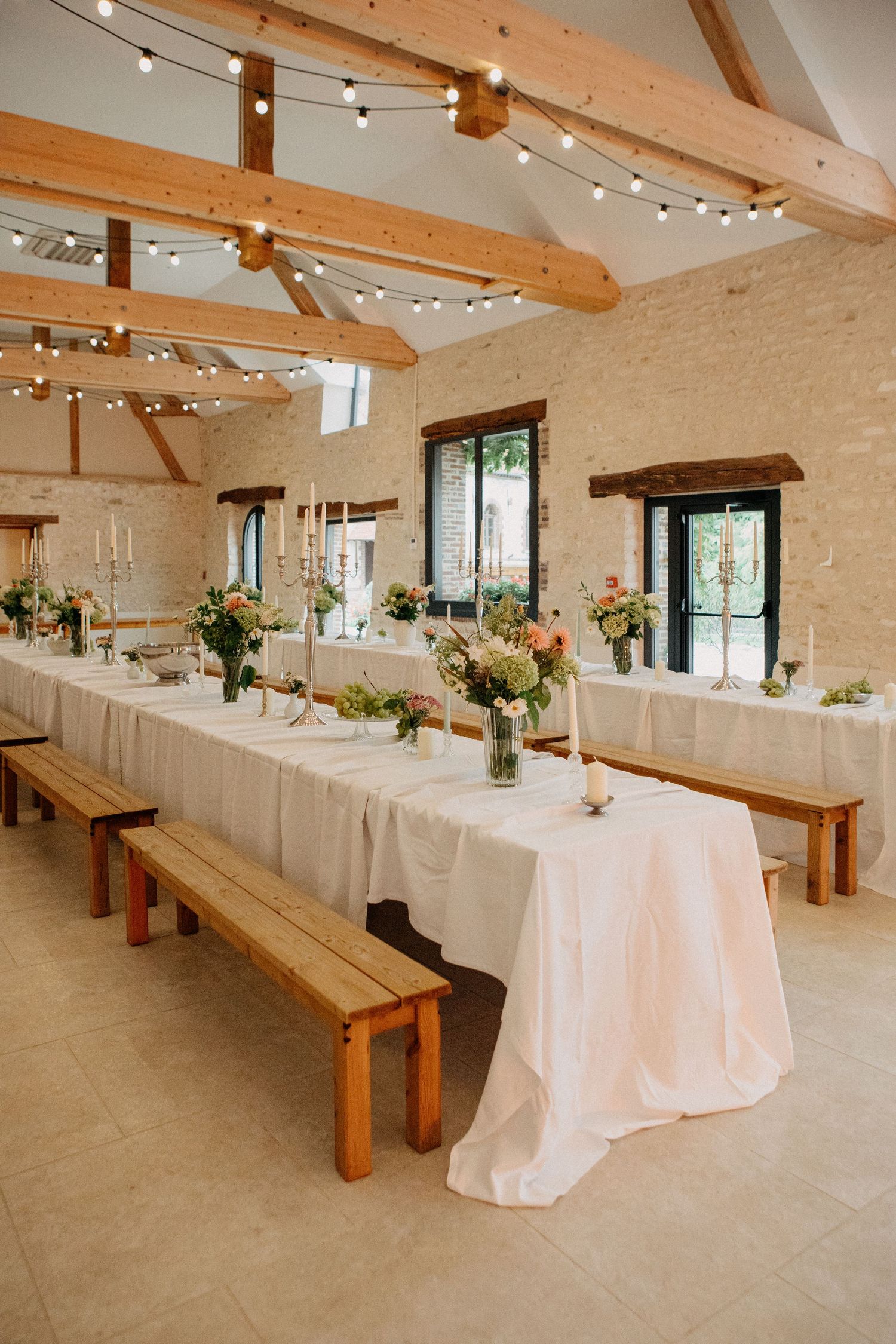 Bright barn wedding reception with exposed beams, string lights, long tables, wooden benches, and floral centerpieces.