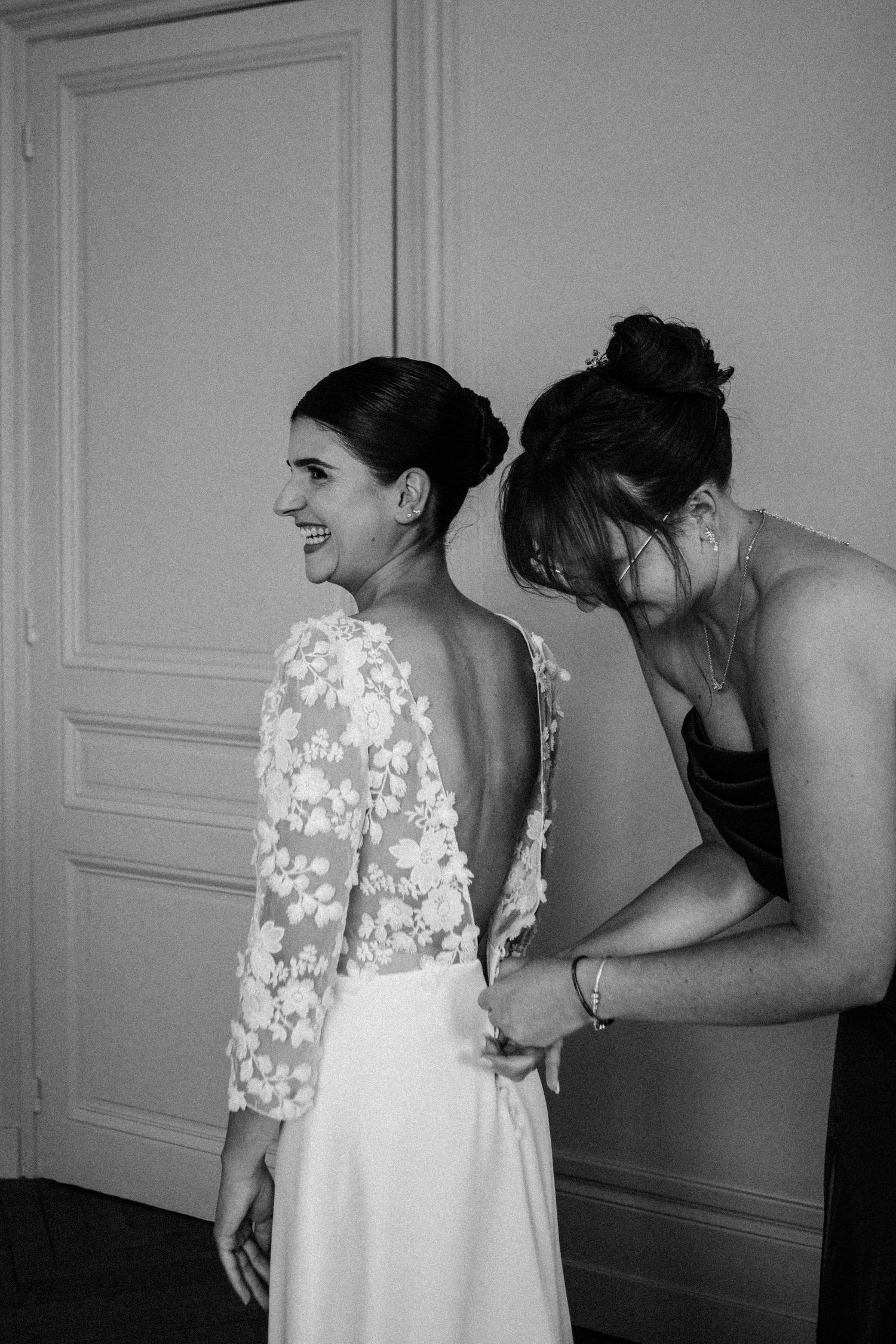 Black and white photo of smiling bride in backless lace gown being buttoned by bridesmaid during prep.