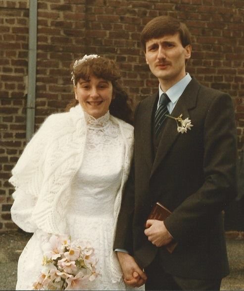 1980s wedding photo of smiling bride in lace dress and groom in dark suit holding hands outside.