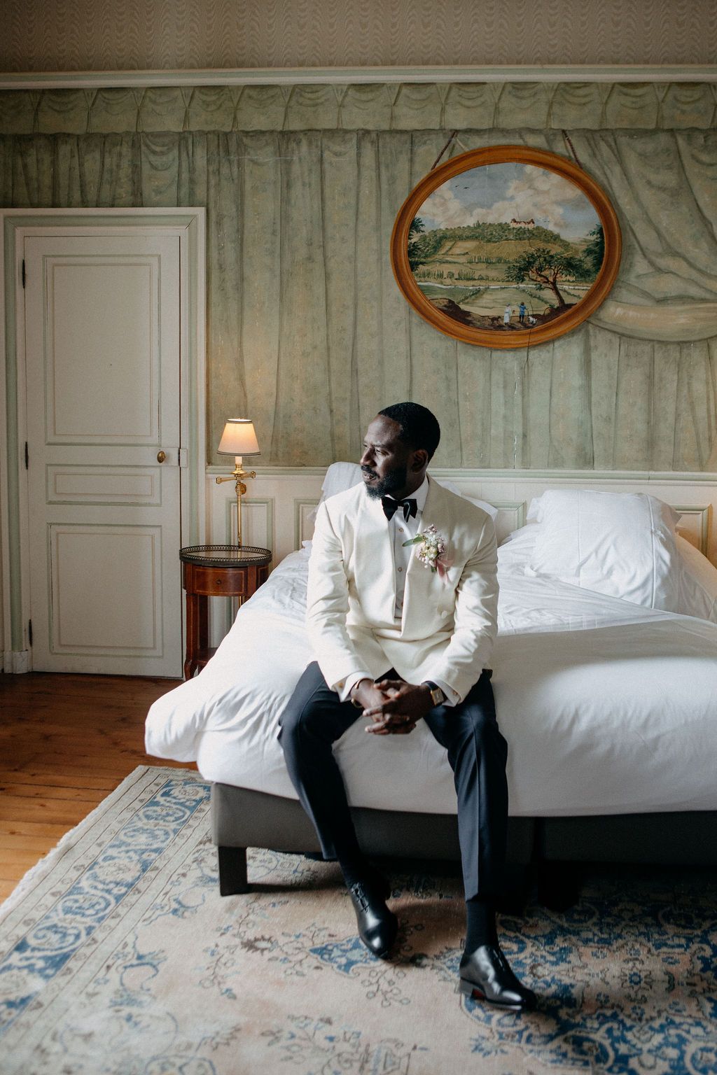 A groom in a cream tuxedo sits on a hotel bed reflecting quietly before his wedding ceremony.