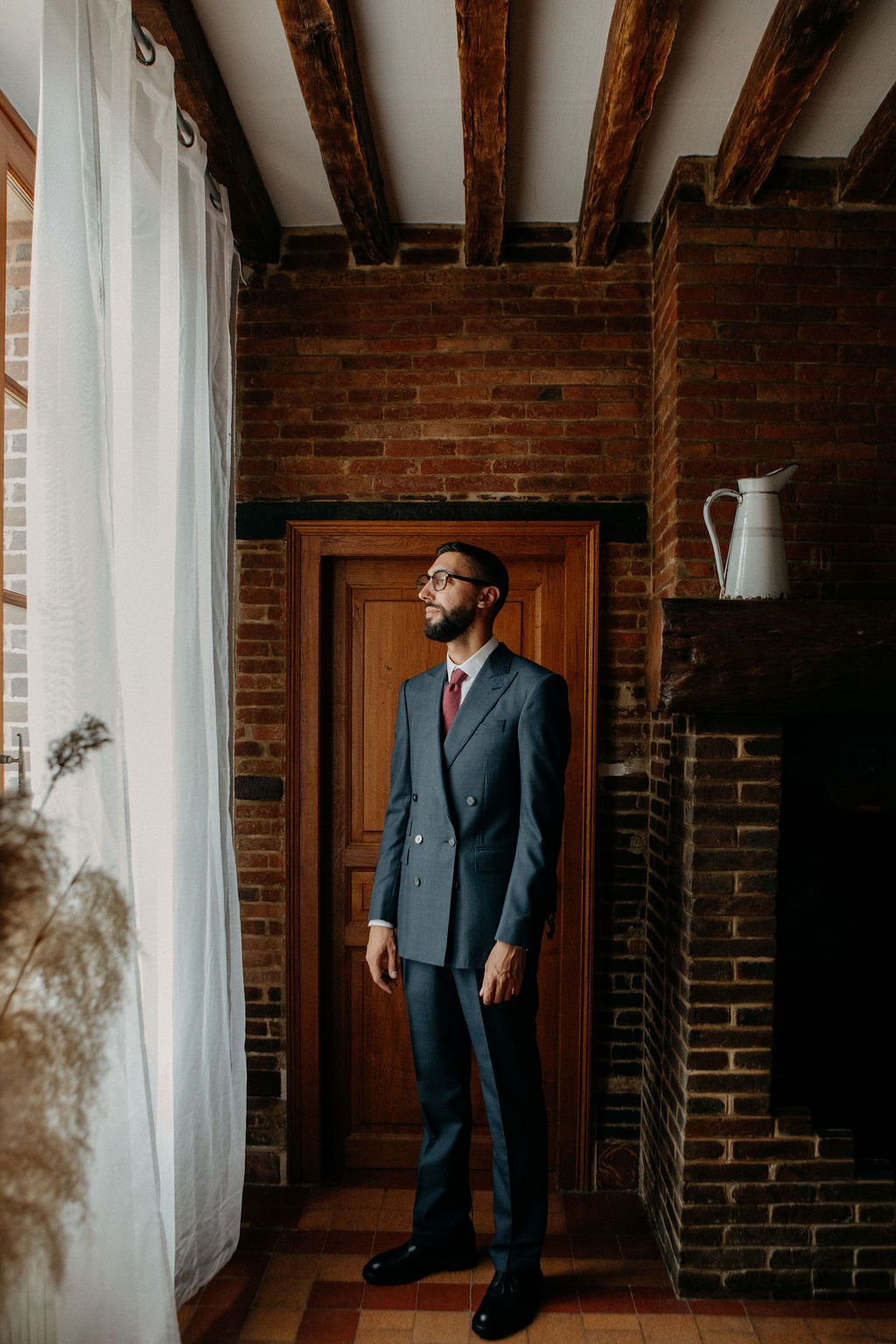 Groom in grey double-breasted suit standing by window in rustic brick interior.