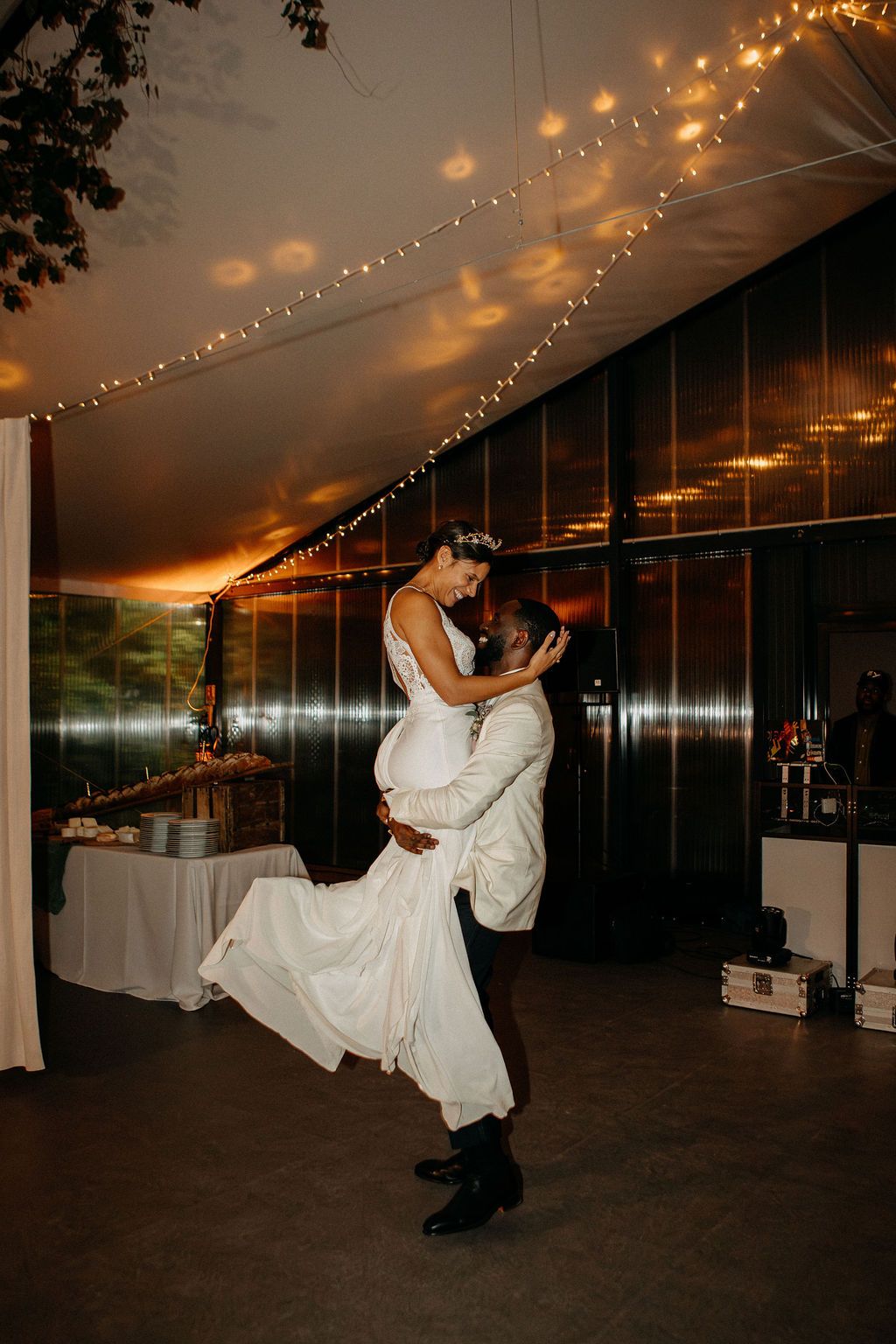 Groom lifting bride during first dance under twinkling fairy lights at romantic wedding reception.
