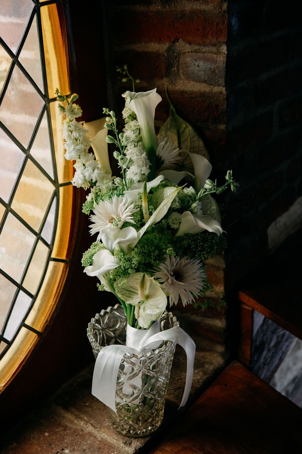 Elegant white floral arrangement with calla lilies and gerberas in crystal vase by stained window.