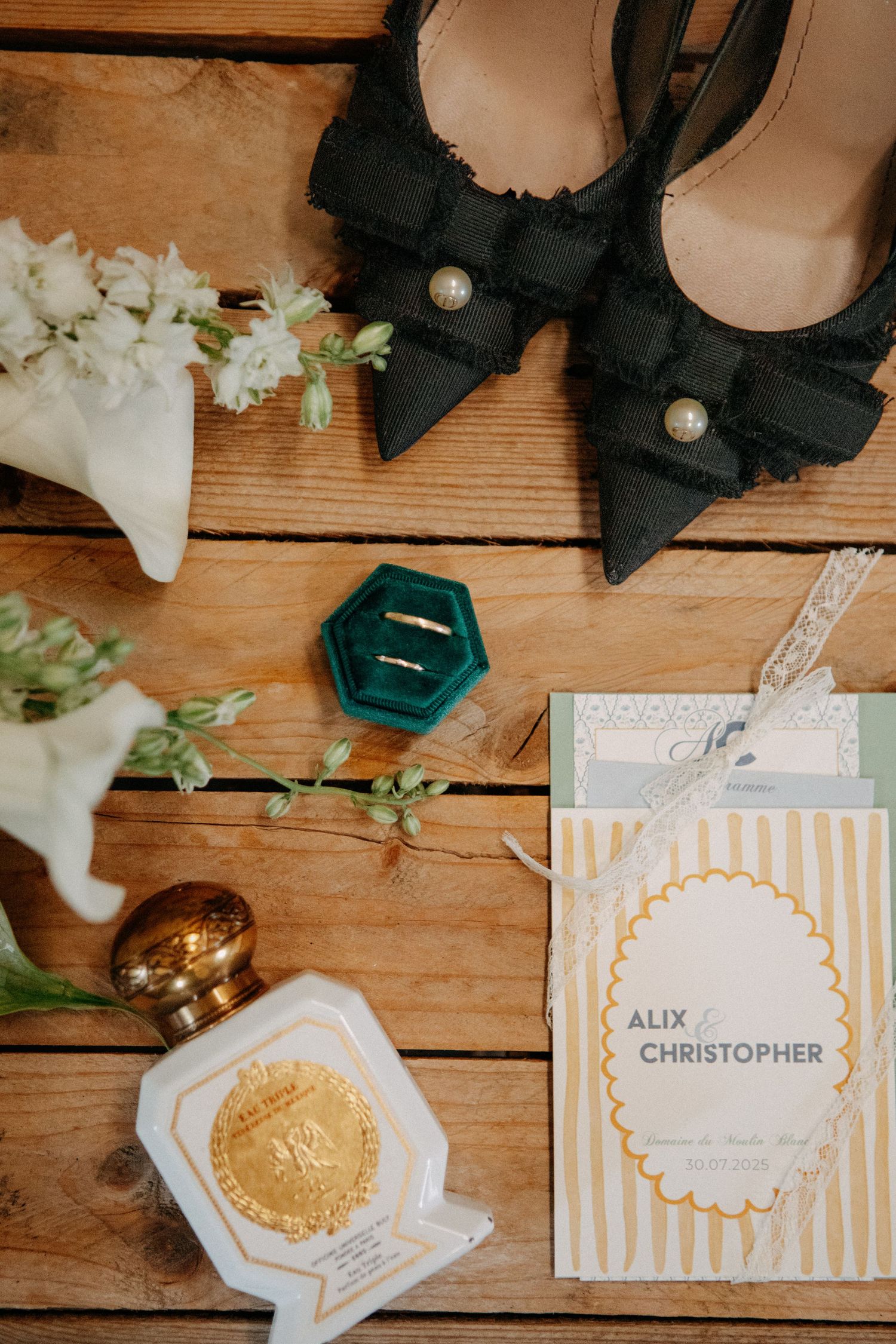 Wedding flat lay featuring black heels, teal ring box, gold rings, perfume, and invitation.