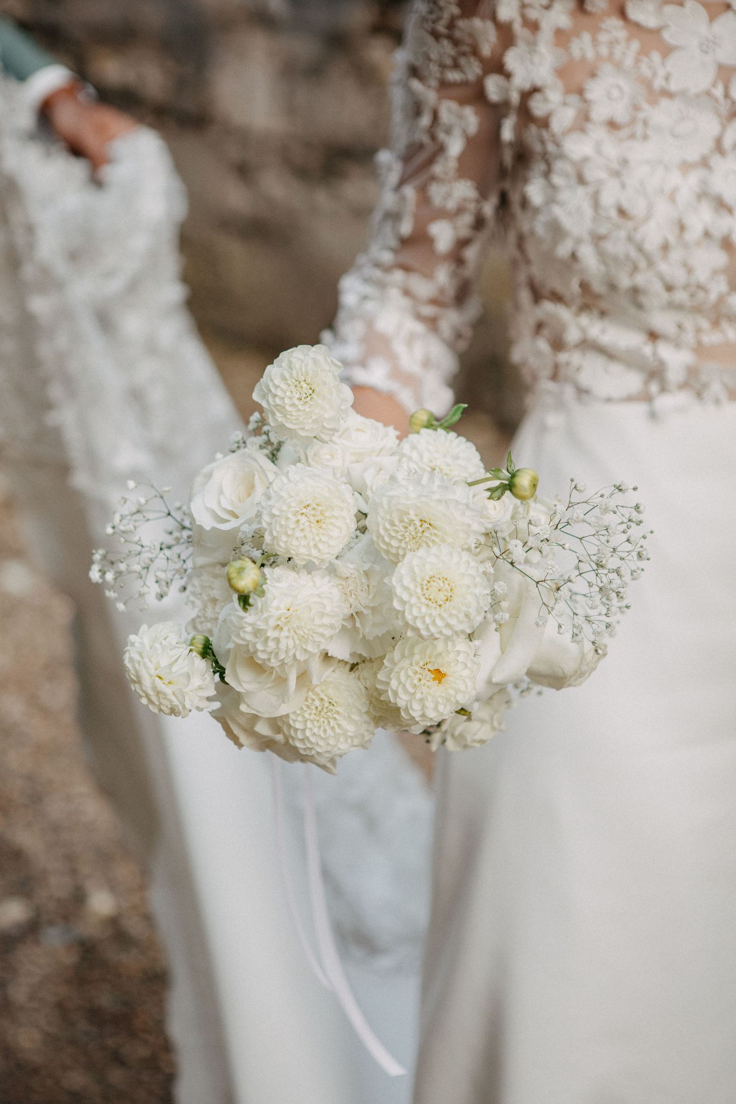 Bride holding elegant white dahlia and baby's breath wedding bouquet against lace gown.