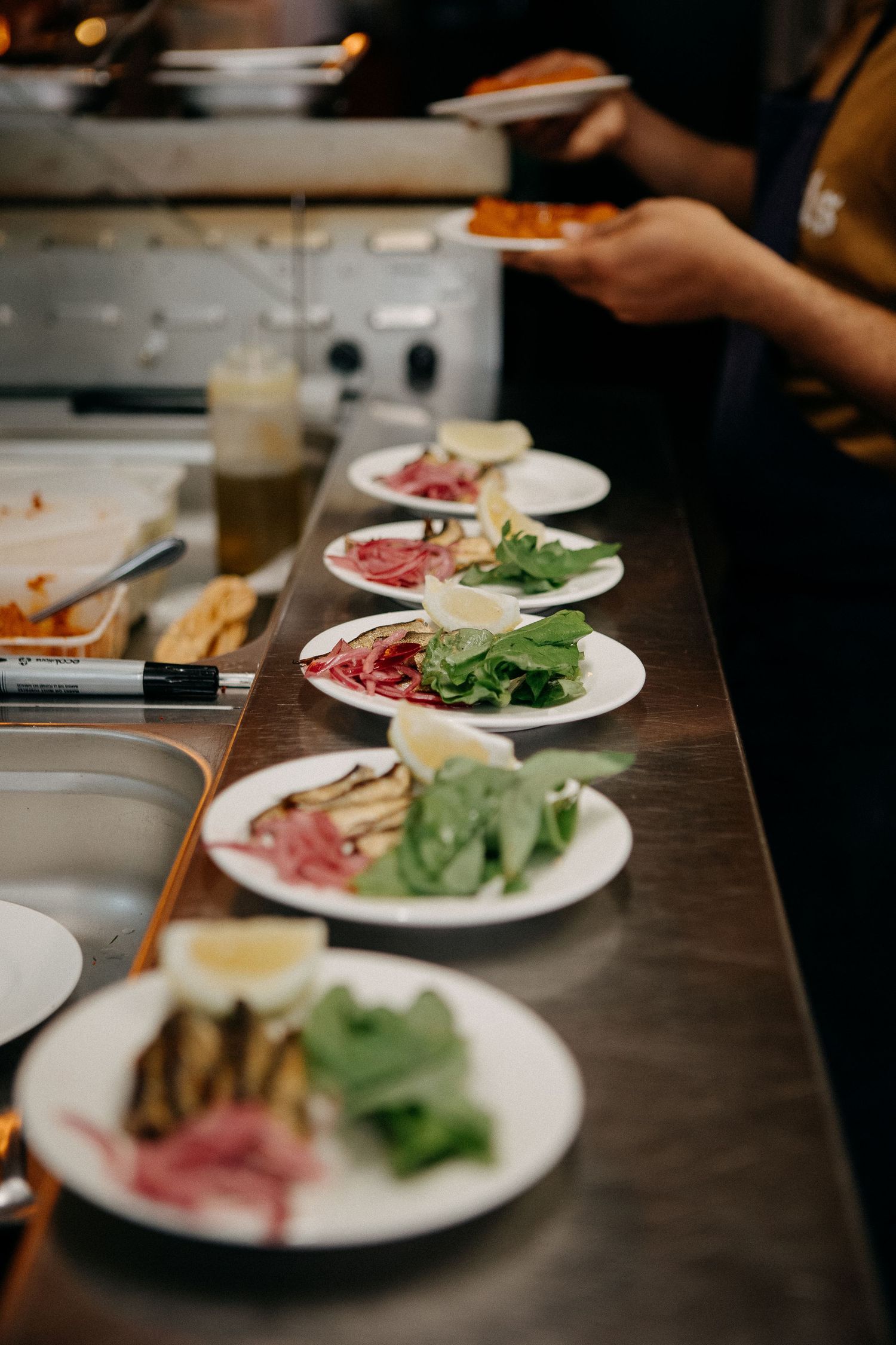 Chef plating fresh salads with spinach and pickled onions on white plates along a restaurant kitchen counter.