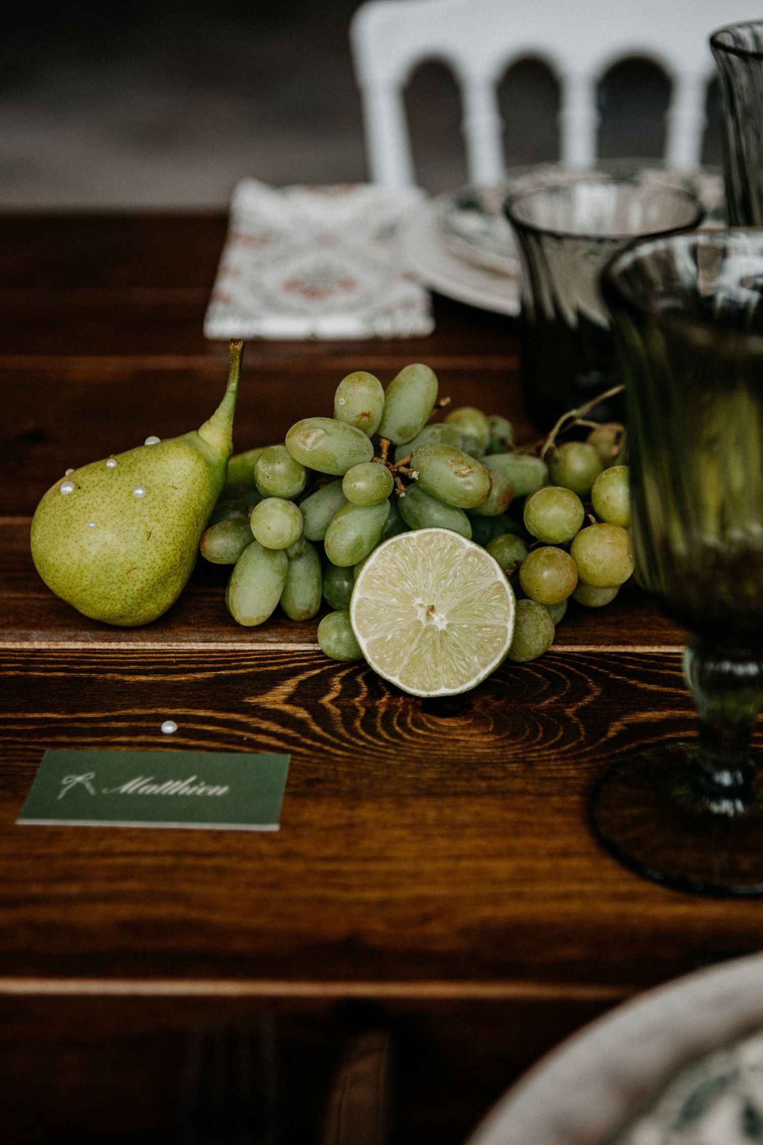 Wedding table setting with green grapes, pear, and halved lime beside a name card on rustic wood.
