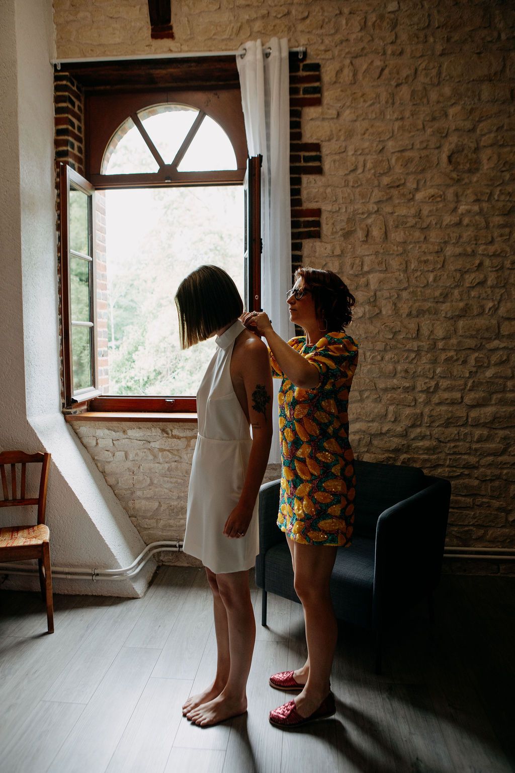 Woman in floral dress helping bride with white halter dress near arched window in stone room.