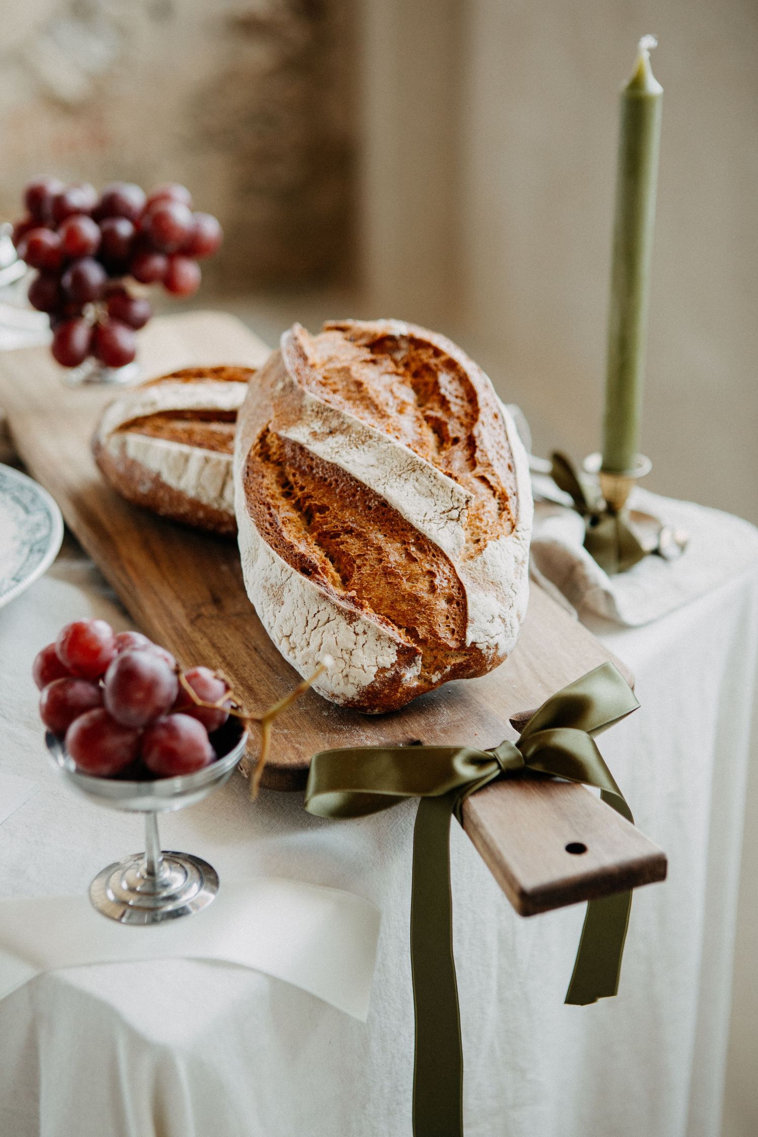 Rustic sourdough bread loaf on a wooden cutting board with red grapes, candle, and green ribbon on a white tablecloth.