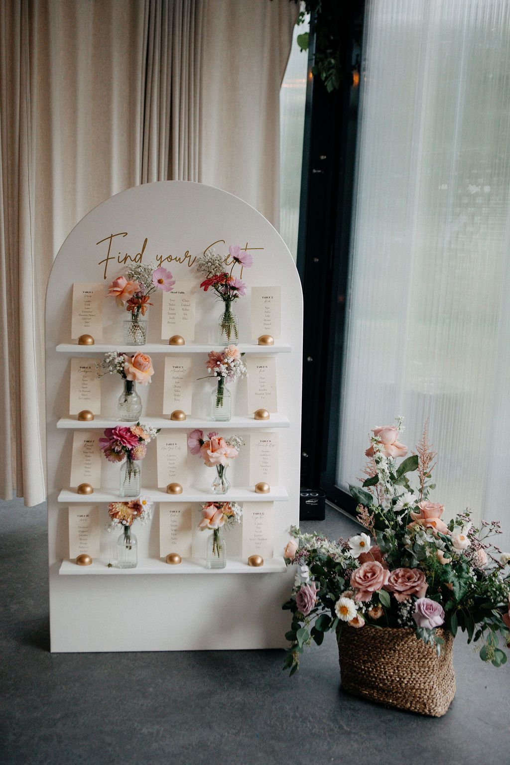 Elegant white arch wedding seating chart display with pink floral arrangements and gold accents.