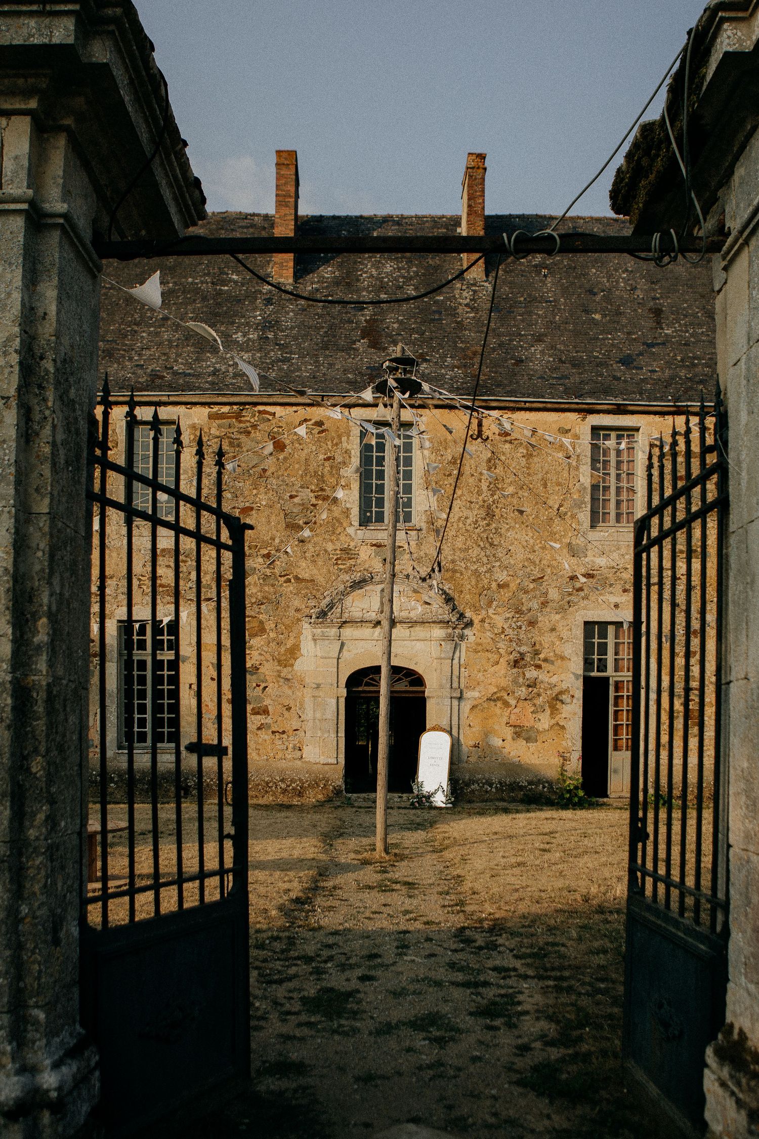 Historic French chateau courtyard viewed through ornate iron gates, featuring aged stone facade and arched doorway.