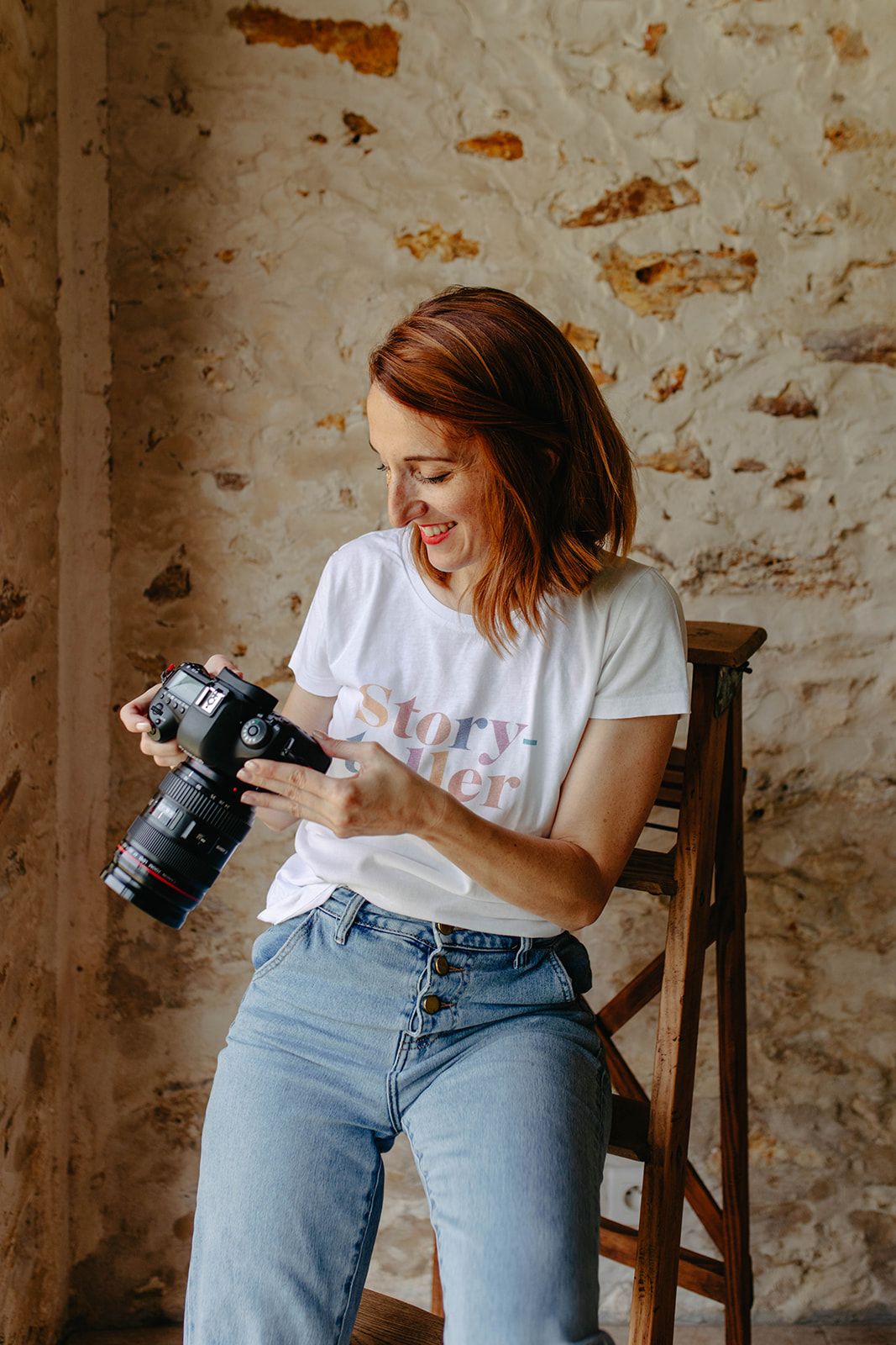 Smiling redhead woman photographer sitting on chair holding DSLR camera wearing Story Teller t-shirt.
