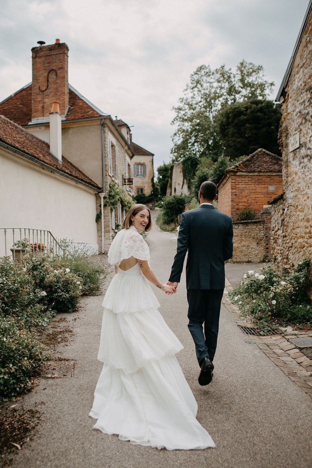 Bride in tiered white gown and groom walk hand in hand through a charming French village pathway.