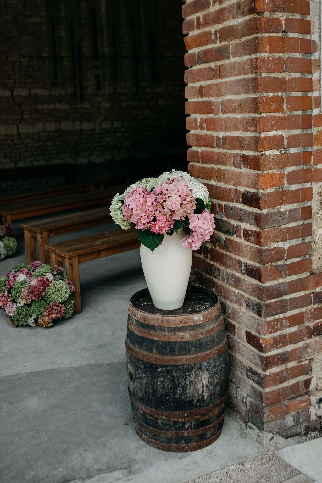 Pink hydrangeas in white vase atop rustic wooden barrel beside brick wall at wedding venue.