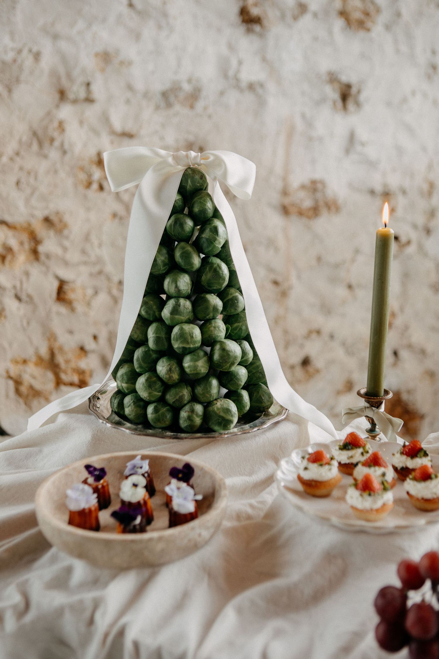 Elegant green croquembouche wedding cake tower with white ribbon bow beside candle and petit fours display.