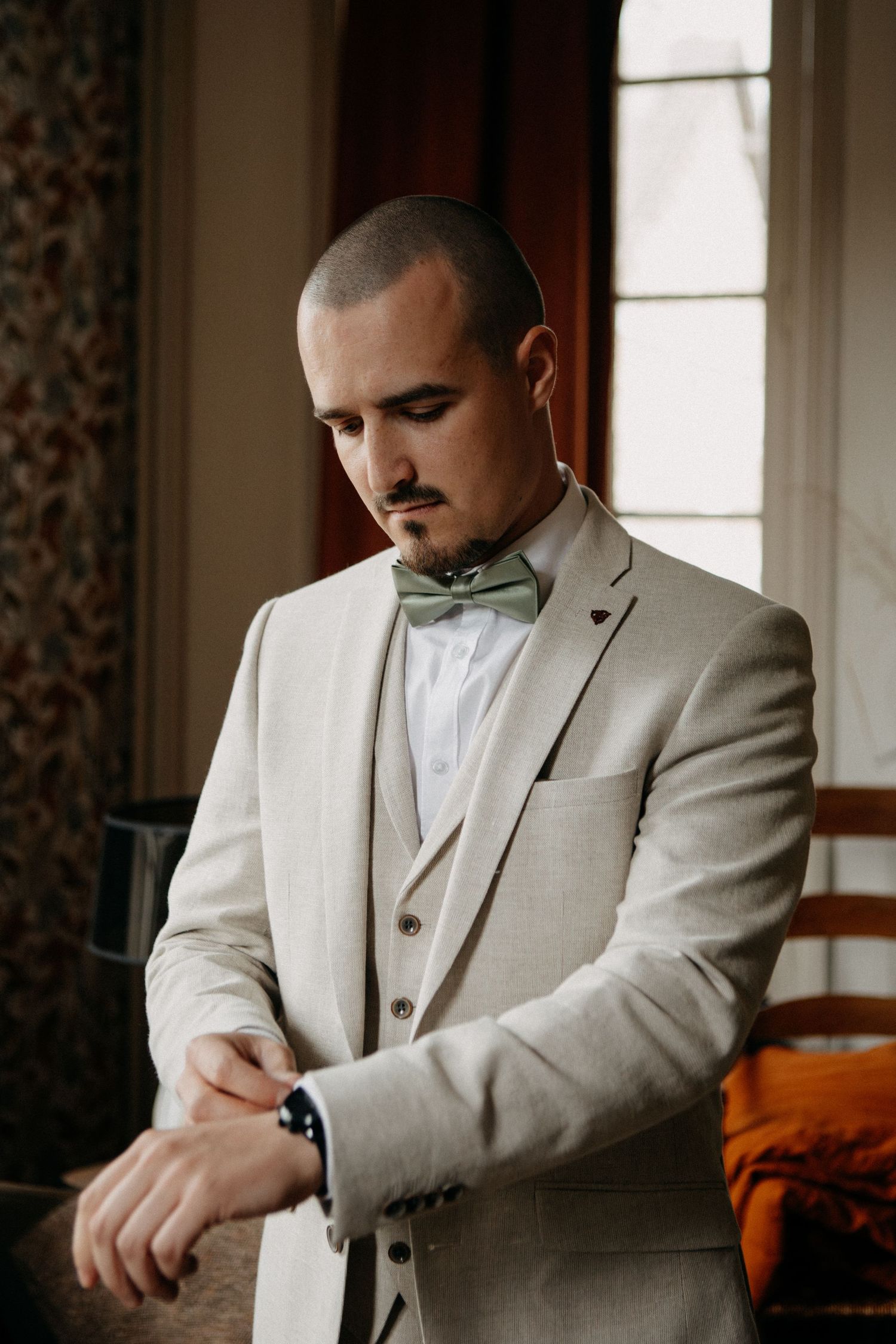 Elegant groom in beige suit with sage green bow tie checking his watch by a window.