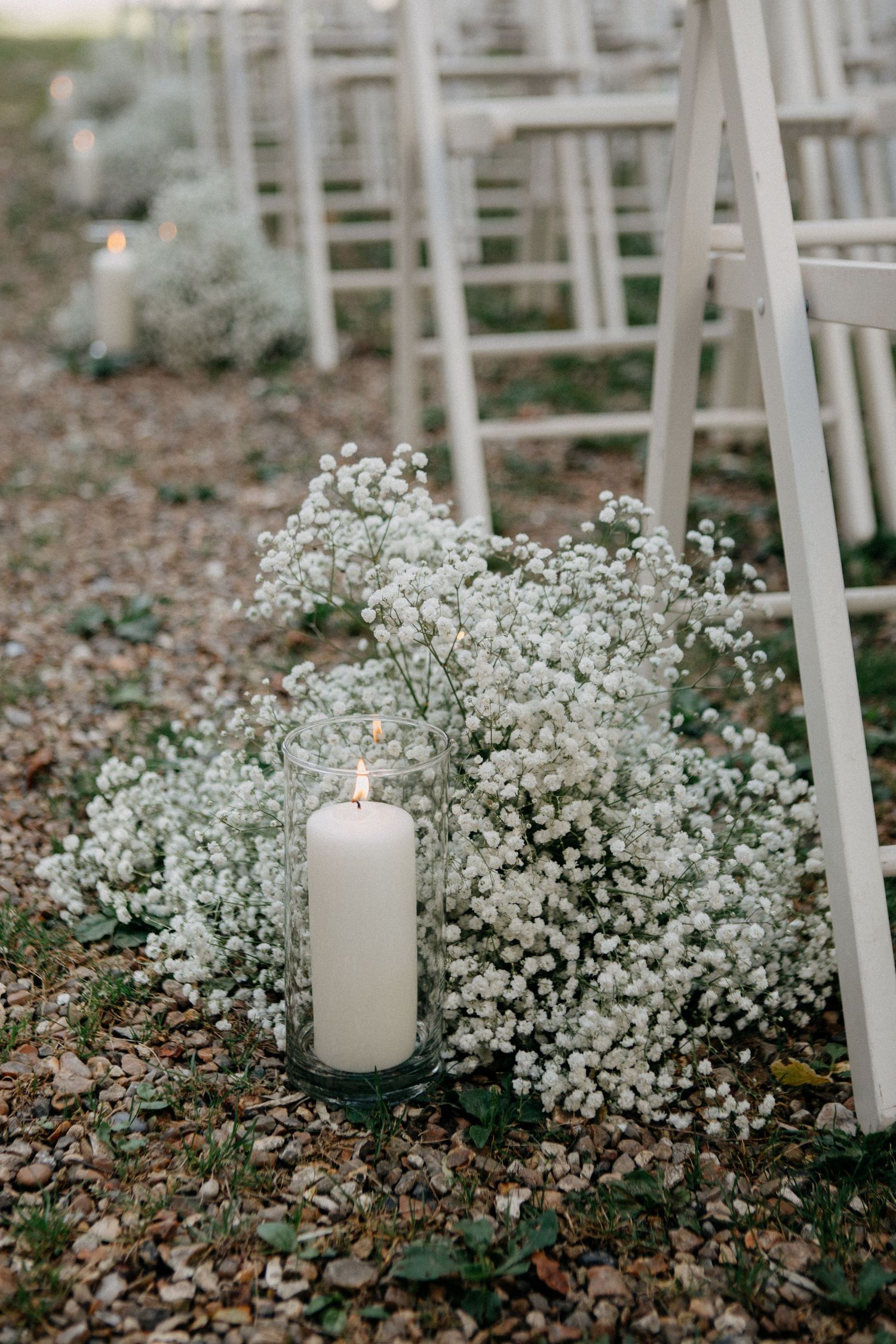 White candles and baby's breath flowers decorating the aisle of an outdoor wedding ceremony with white chairs.