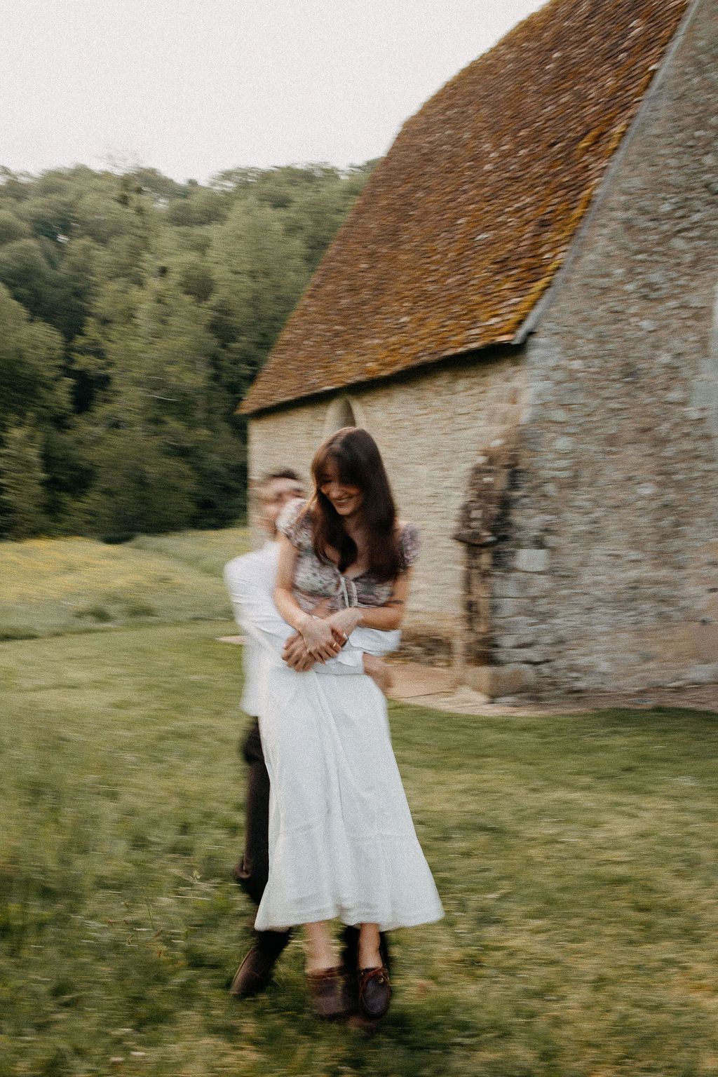 A joyful couple embraces and spins near a historic stone building surrounded by lush greenery.