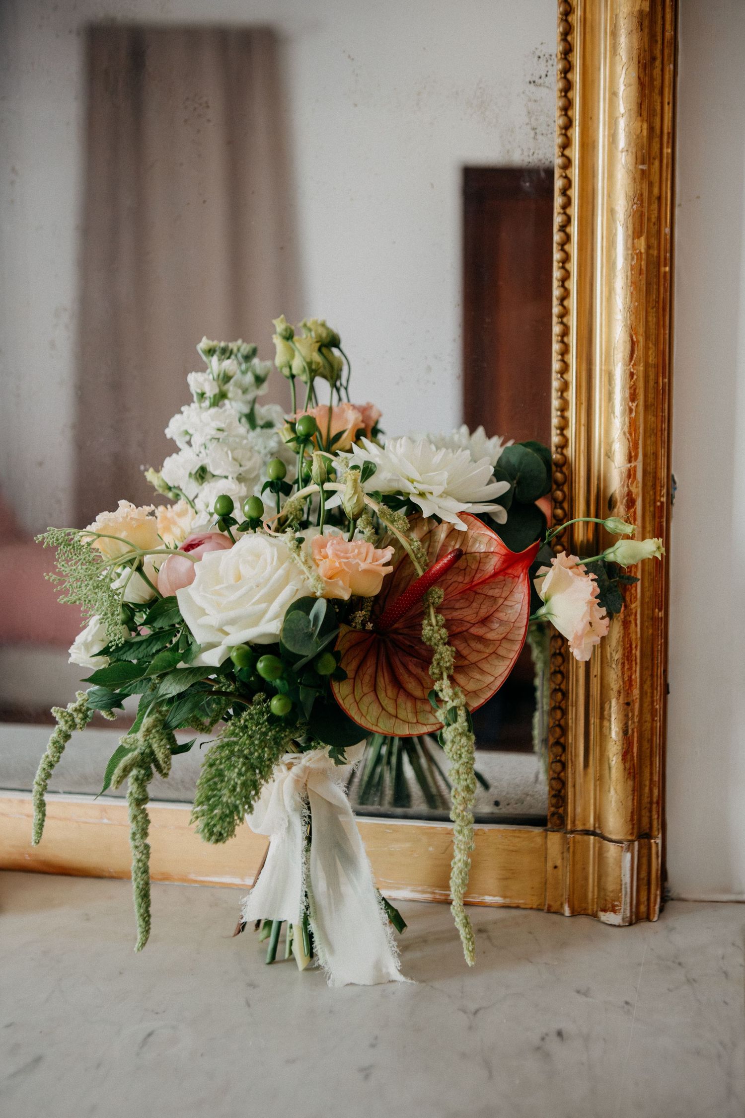 Elegant bridal bouquet with white roses, peach blooms, anthurium, and greenery leaning against a gold ornate mirror.