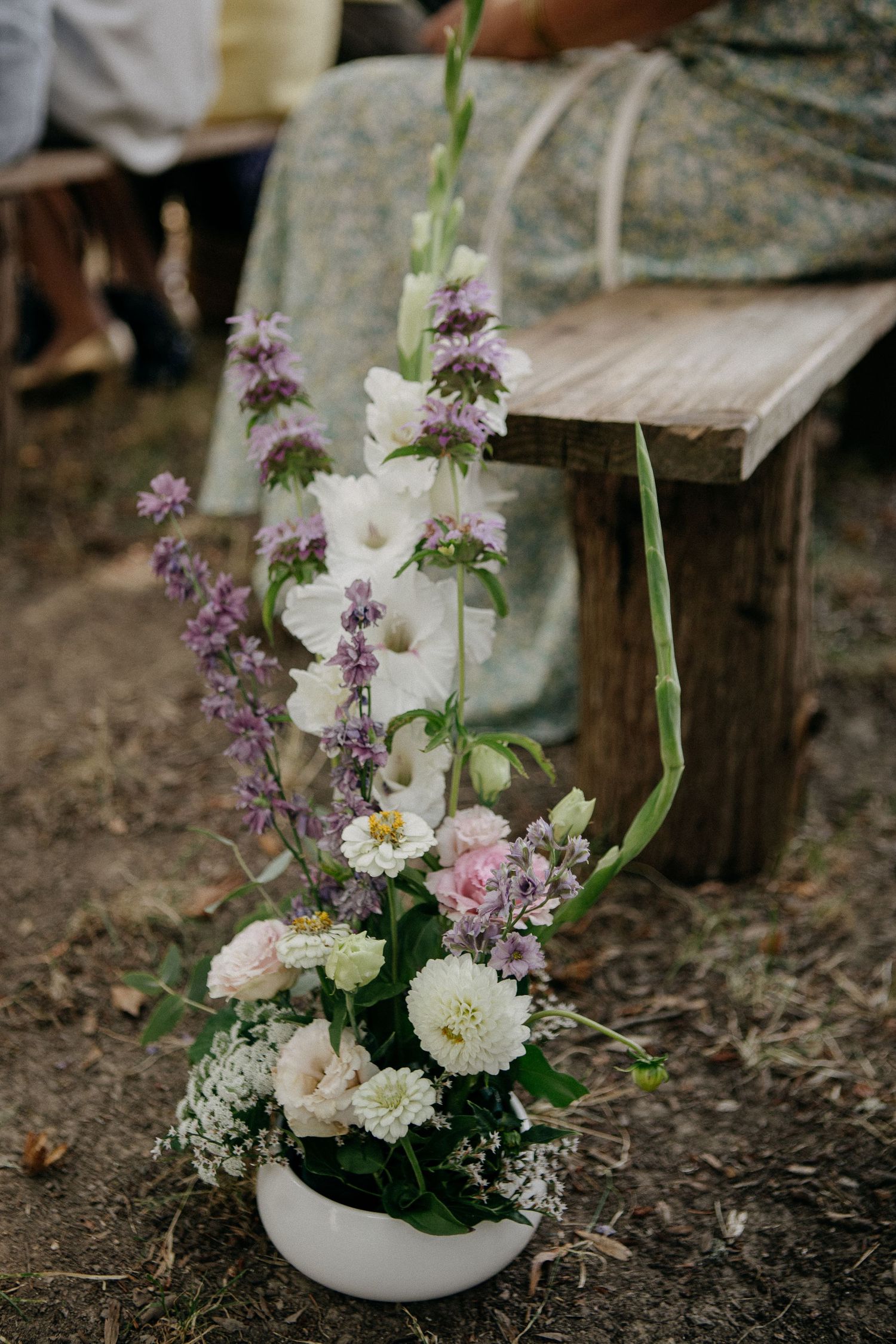 White ceramic vase with wildflowers, gladiolus, and baby's breath beside a rustic wooden bench at an outdoor wedding.