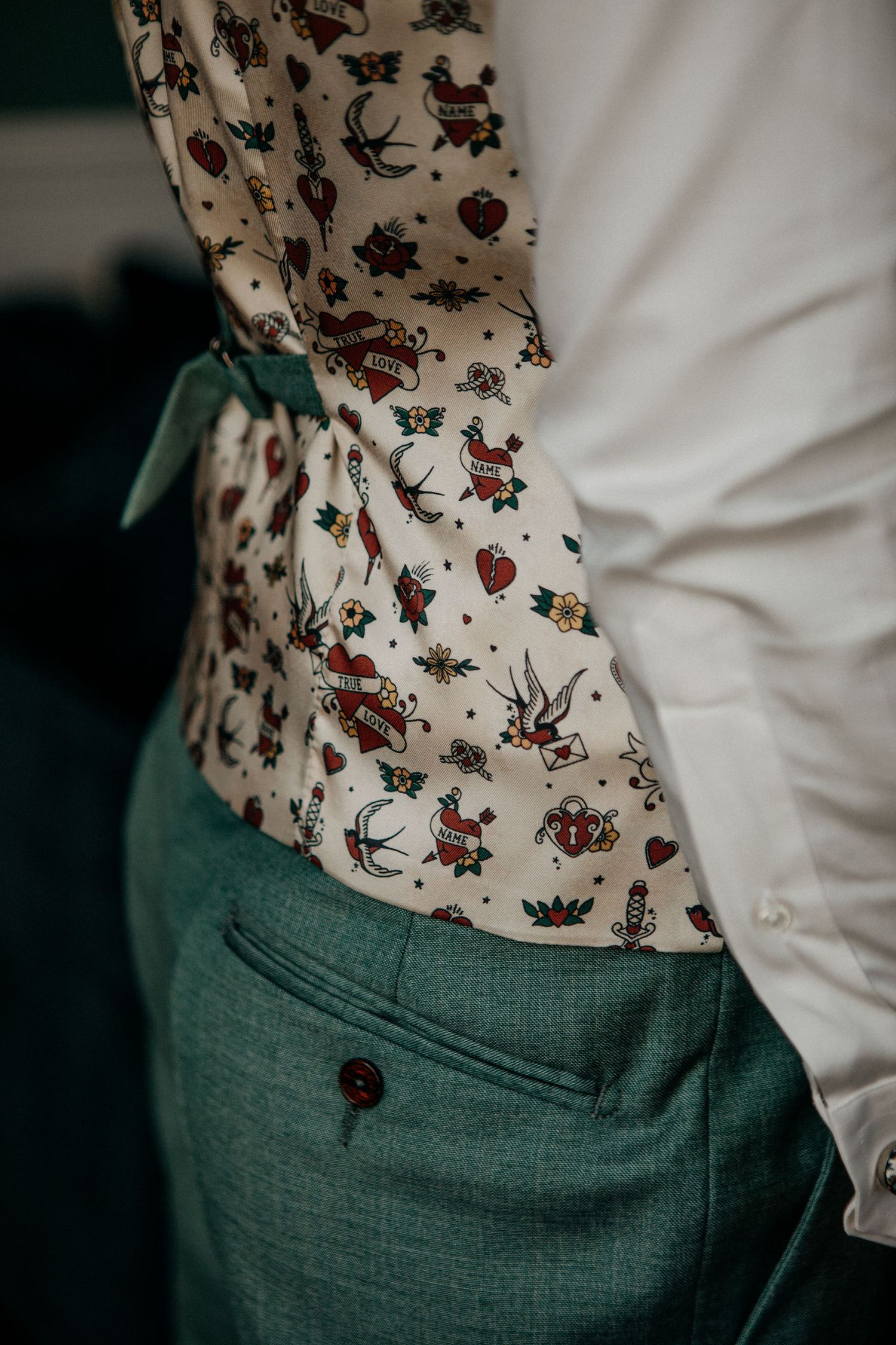 Close-up detail of groom's colorful tattoo-print patterned vest with green trousers during wedding preparations.