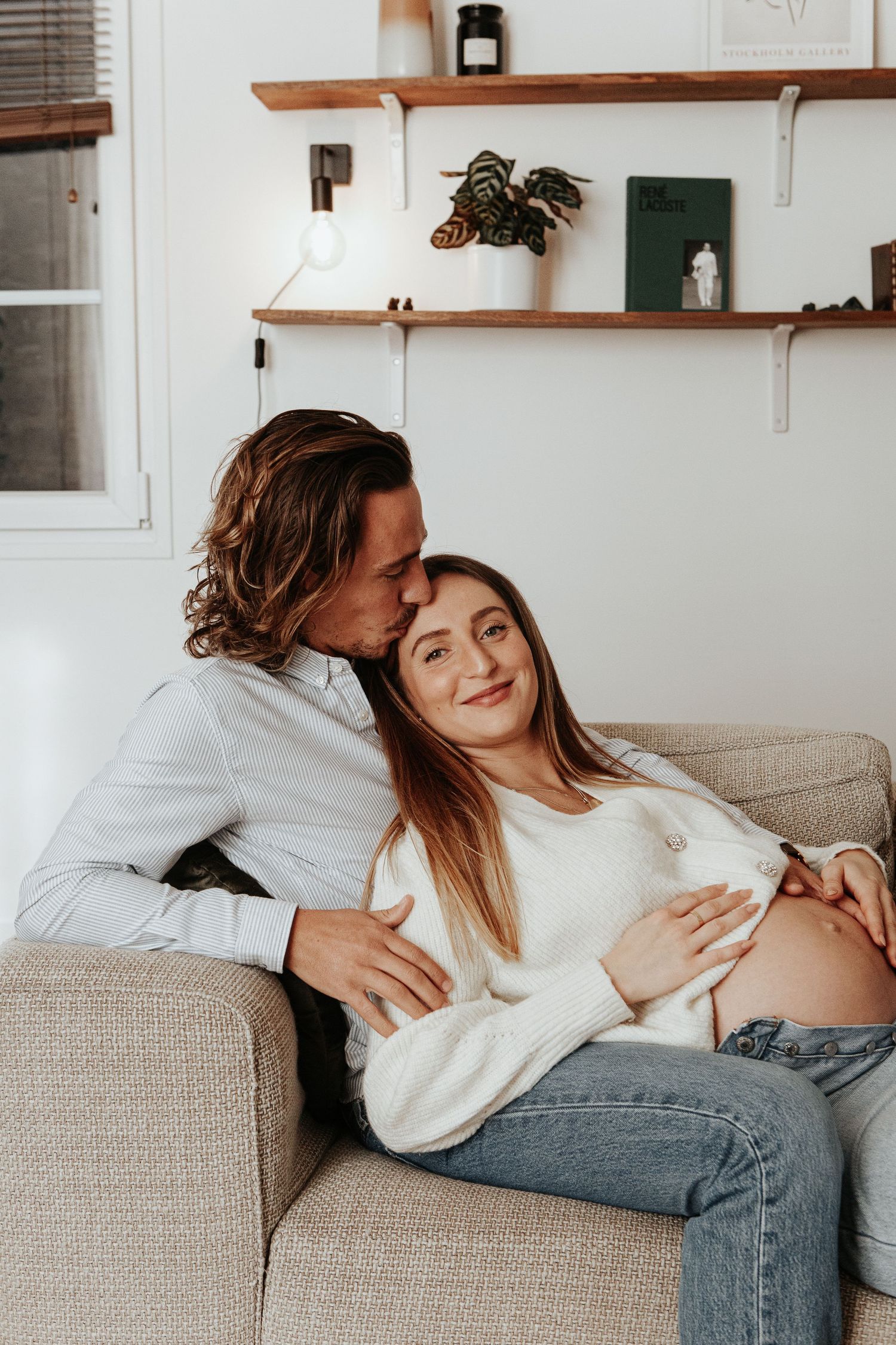 Man kissing pregnant woman's head while embracing her on couch in cozy home setting.