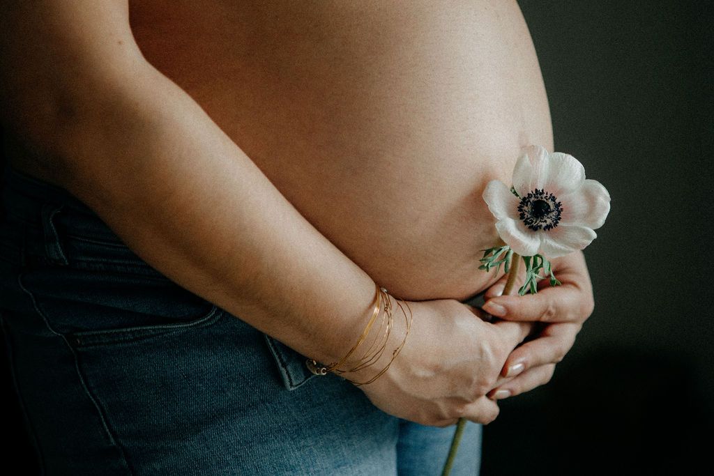 Close-up of pregnant woman's baby bump holding a single white anemone flower, wearing jeans and a gold bracelet.