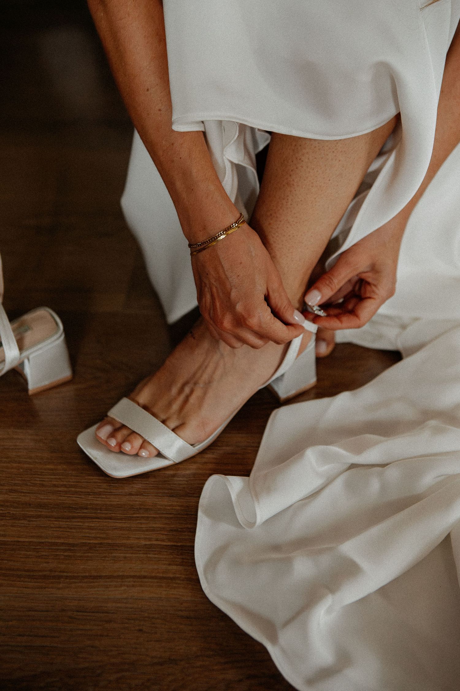 Bride fastening white block heel sandal straps on wedding day, wearing gold bracelet and flowing white bridal gown.