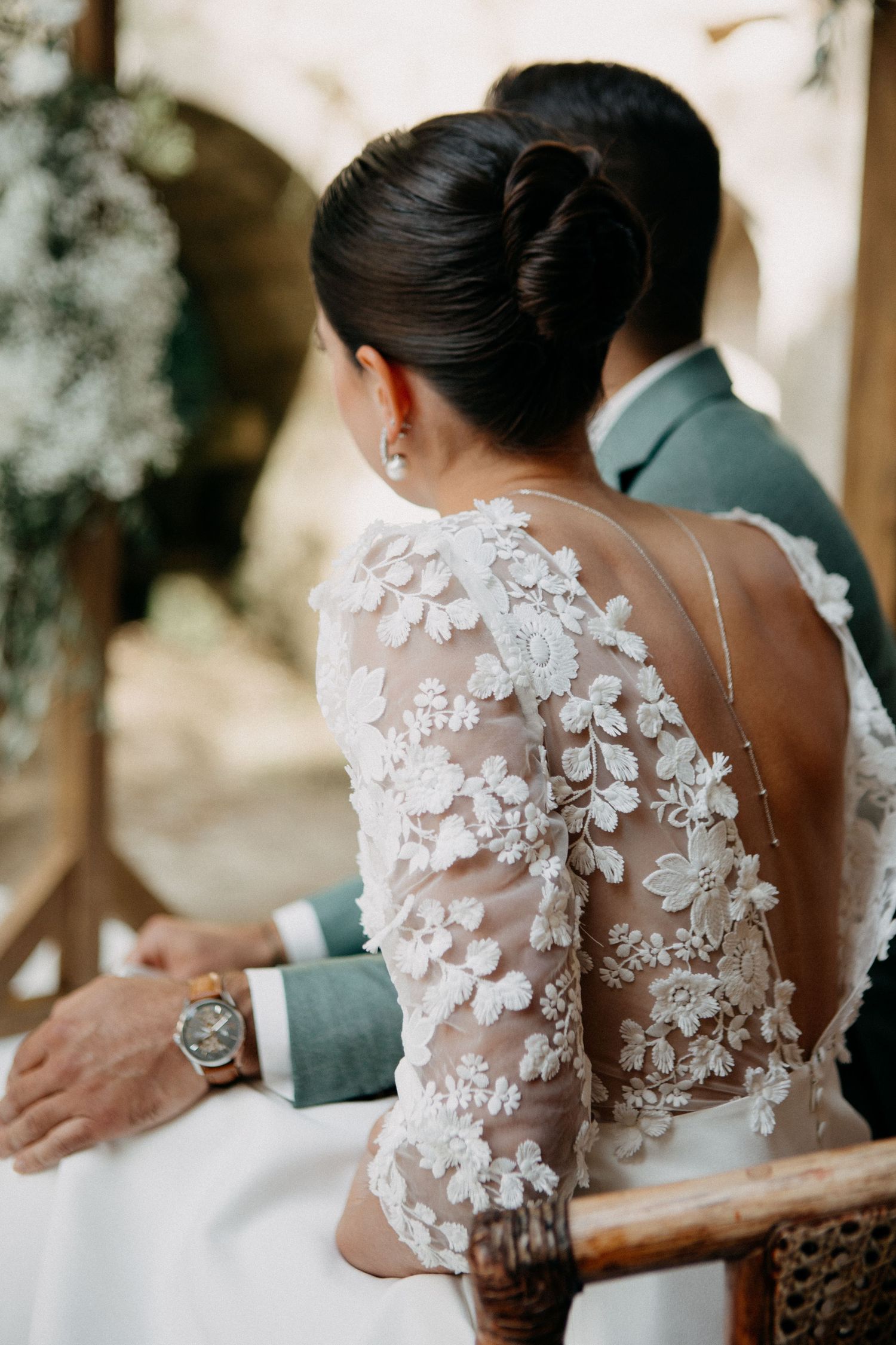 Close-up of bride's floral lace long-sleeve open-back gown and elegant updo beside groom in sage suit.