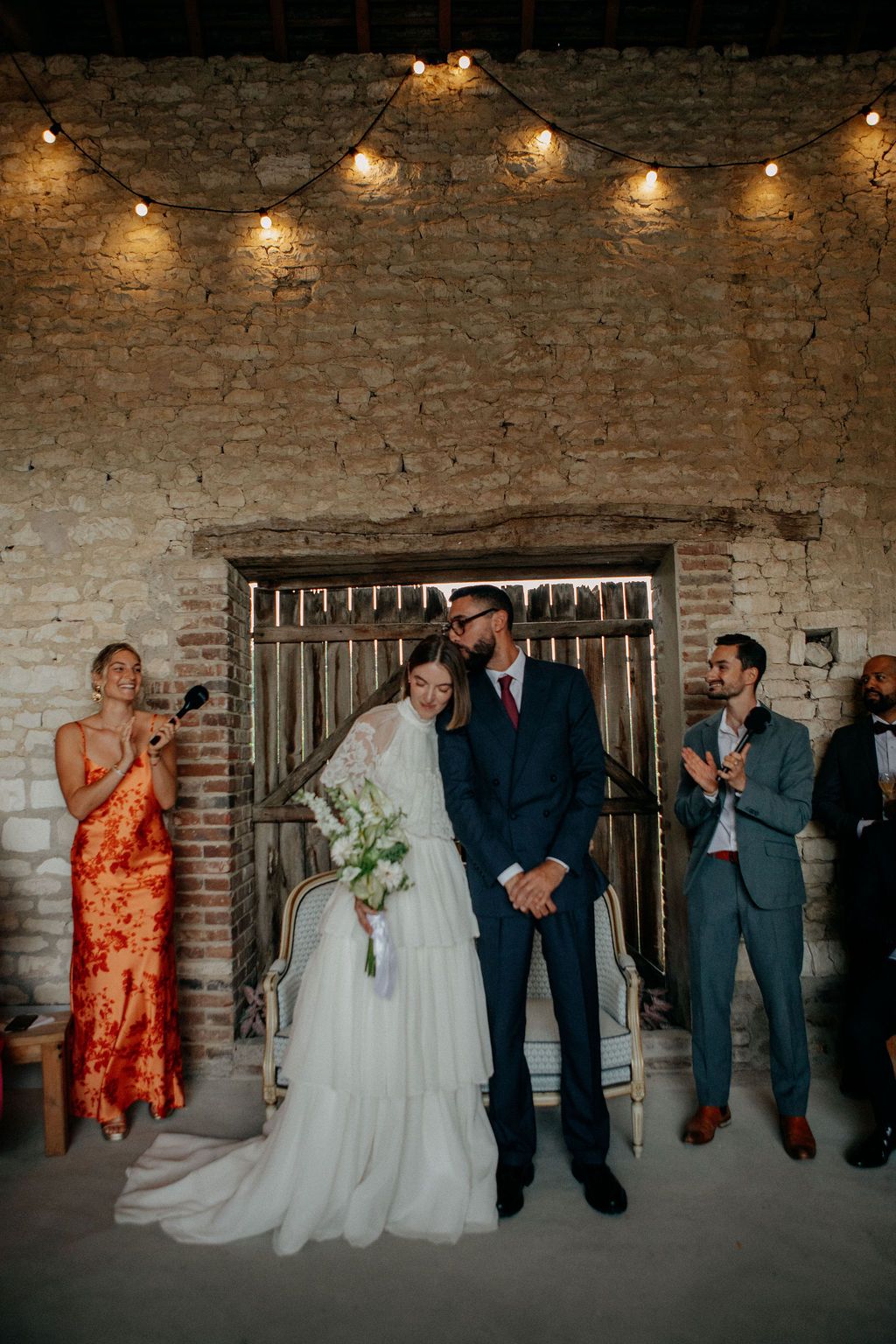 Newlyweds share a kiss under warm string lights in a rustic stone barn wedding venue.