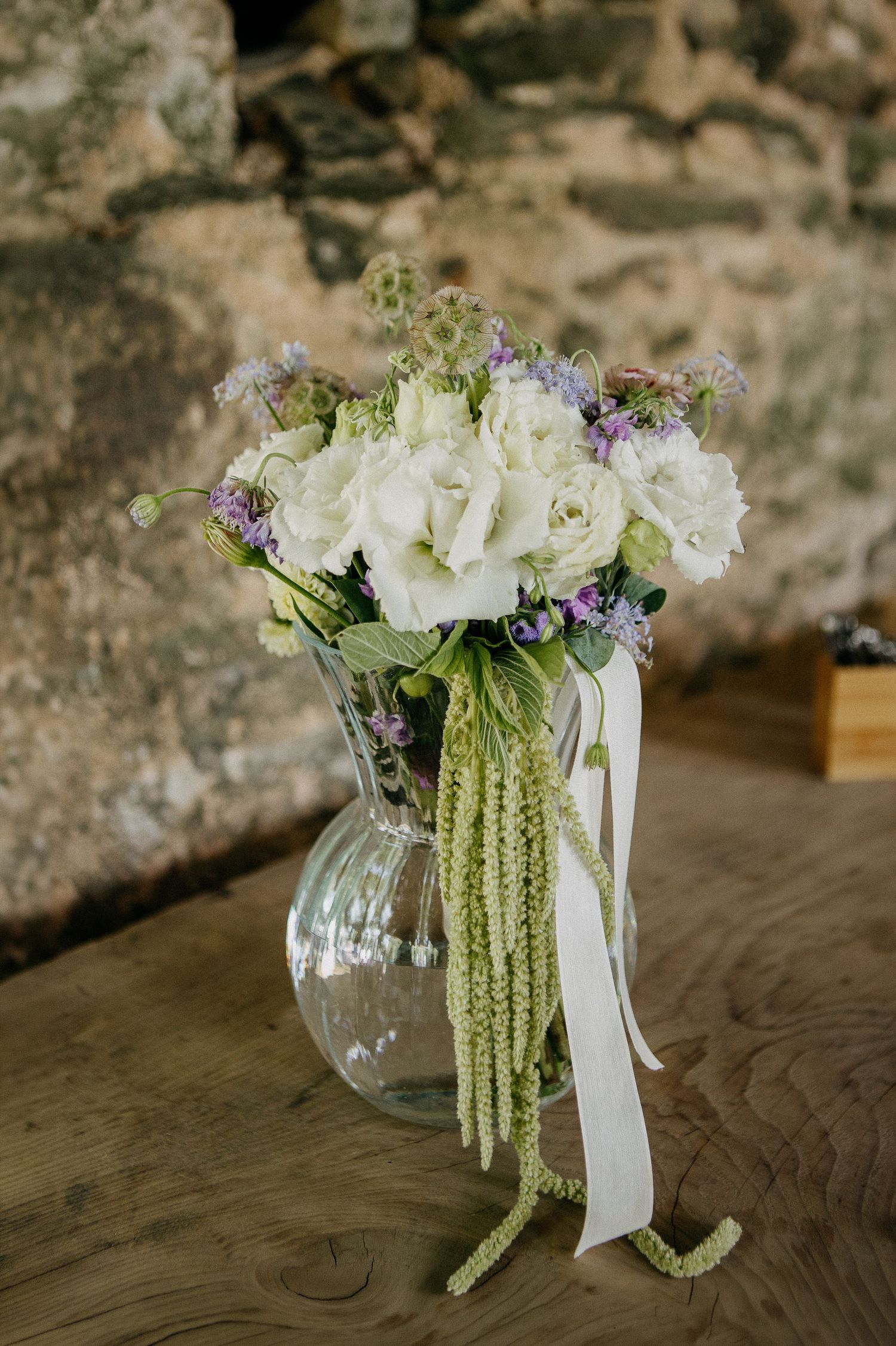 Elegant wedding bouquet with white roses and purple wildflowers in a glass vase, adorned with green trailing amaranthus and white ribbon.