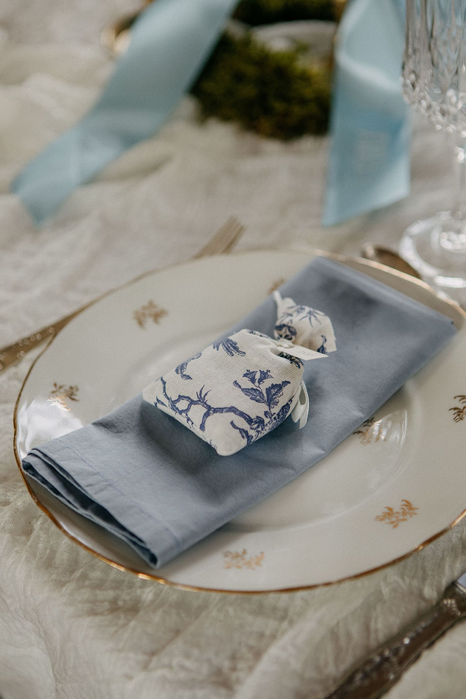 Elegant wedding place setting with blue linen napkin, floral favor, gold-rimmed china plate, and light blue ribbon accent.