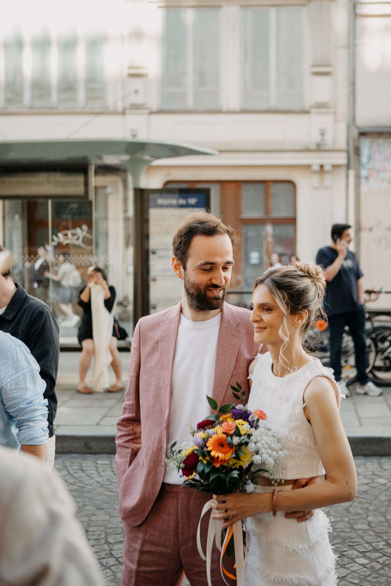 Bride and groom smiling together outdoors, she holds a colorful bouquet, groom in pink suit, urban street background.