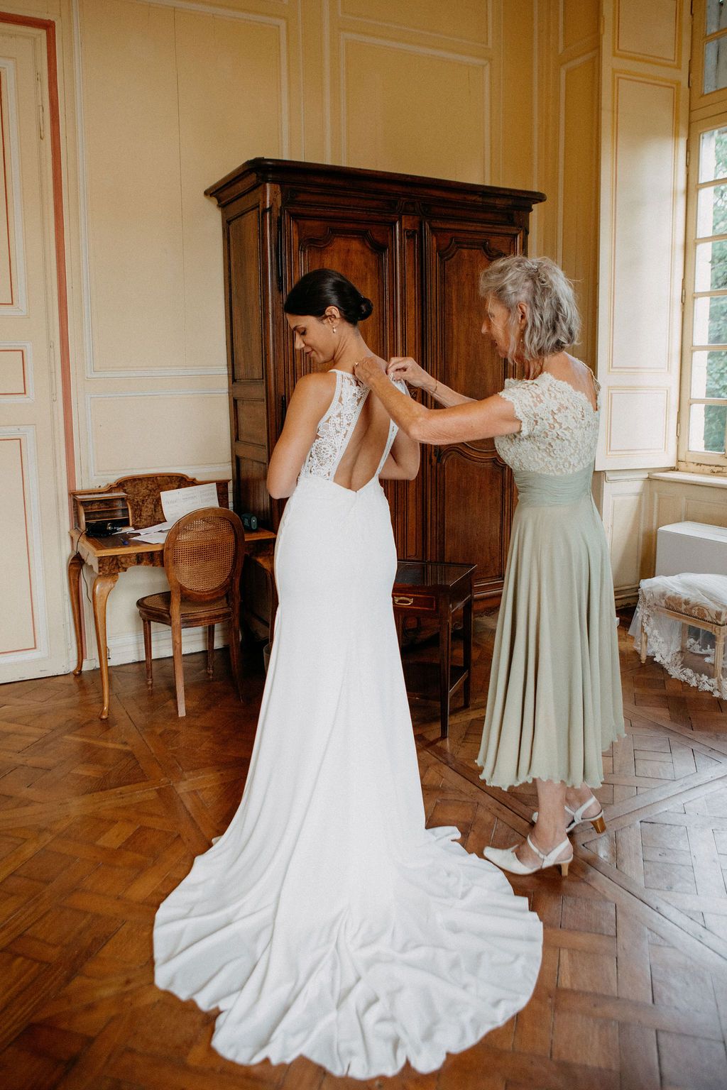 Bride in white lace-back wedding gown being helped by older woman in sage green dress in elegant room.