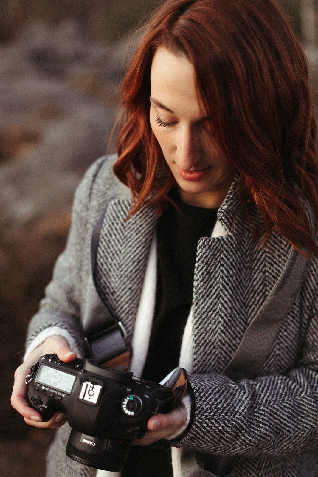 Red-haired woman in gray coat reviewing photos on Canon DSLR camera outdoors.