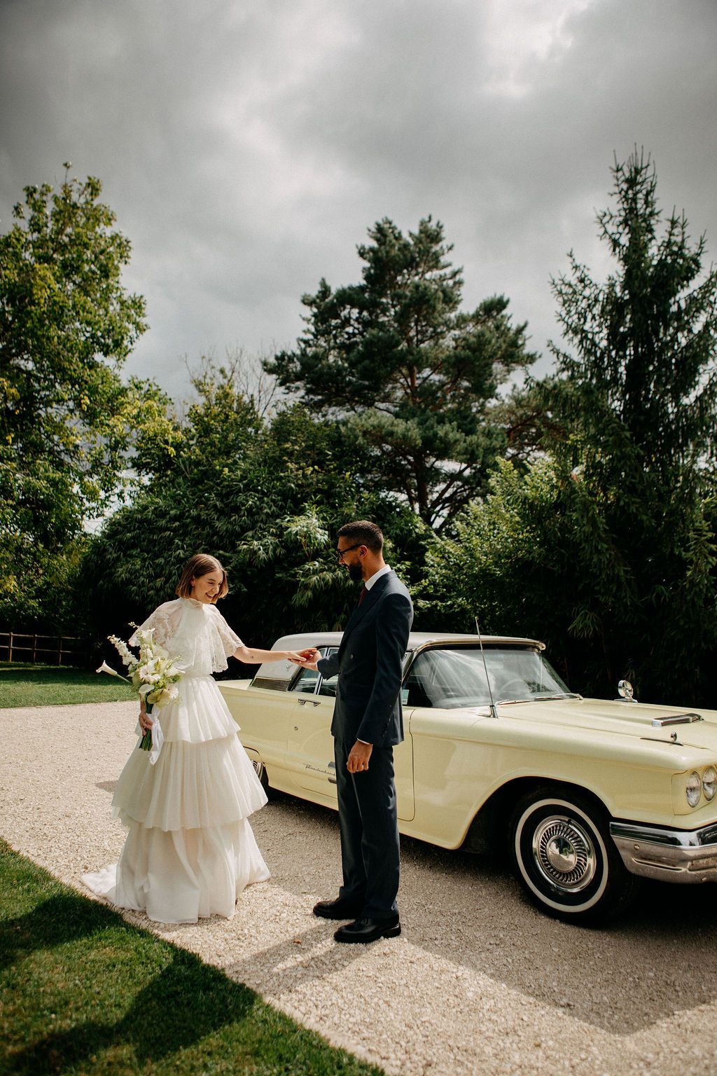 Elegant bride hands keys to groom beside a classic pale yellow convertible on their wedding day.
