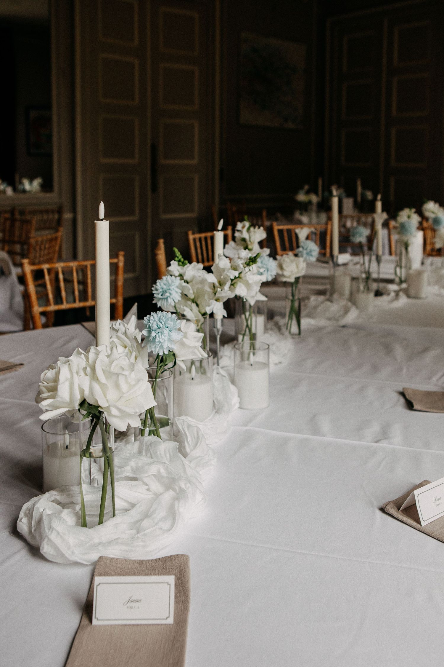 Elegant wedding reception table with white floral centerpieces, candles, and soft blue hydrangeas on a white linen tablecloth.