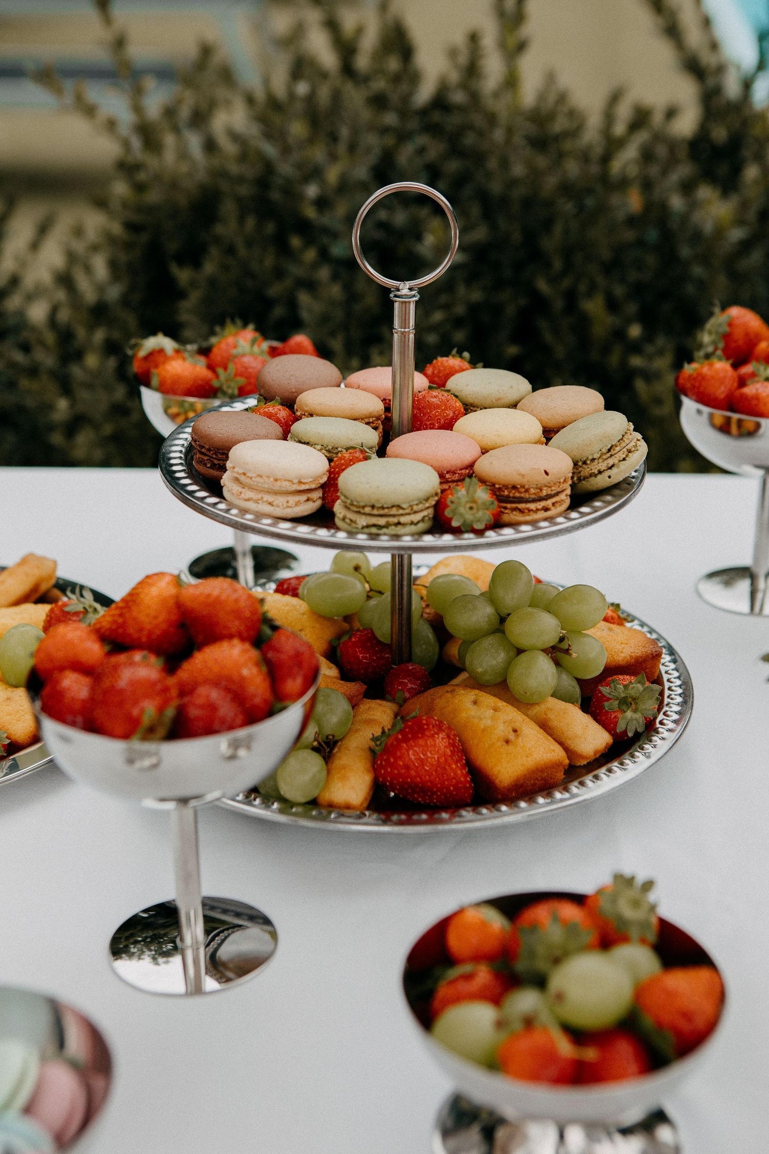 Elegant two-tier silver stand with macarons and fresh fruit including strawberries and grapes at an outdoor event.