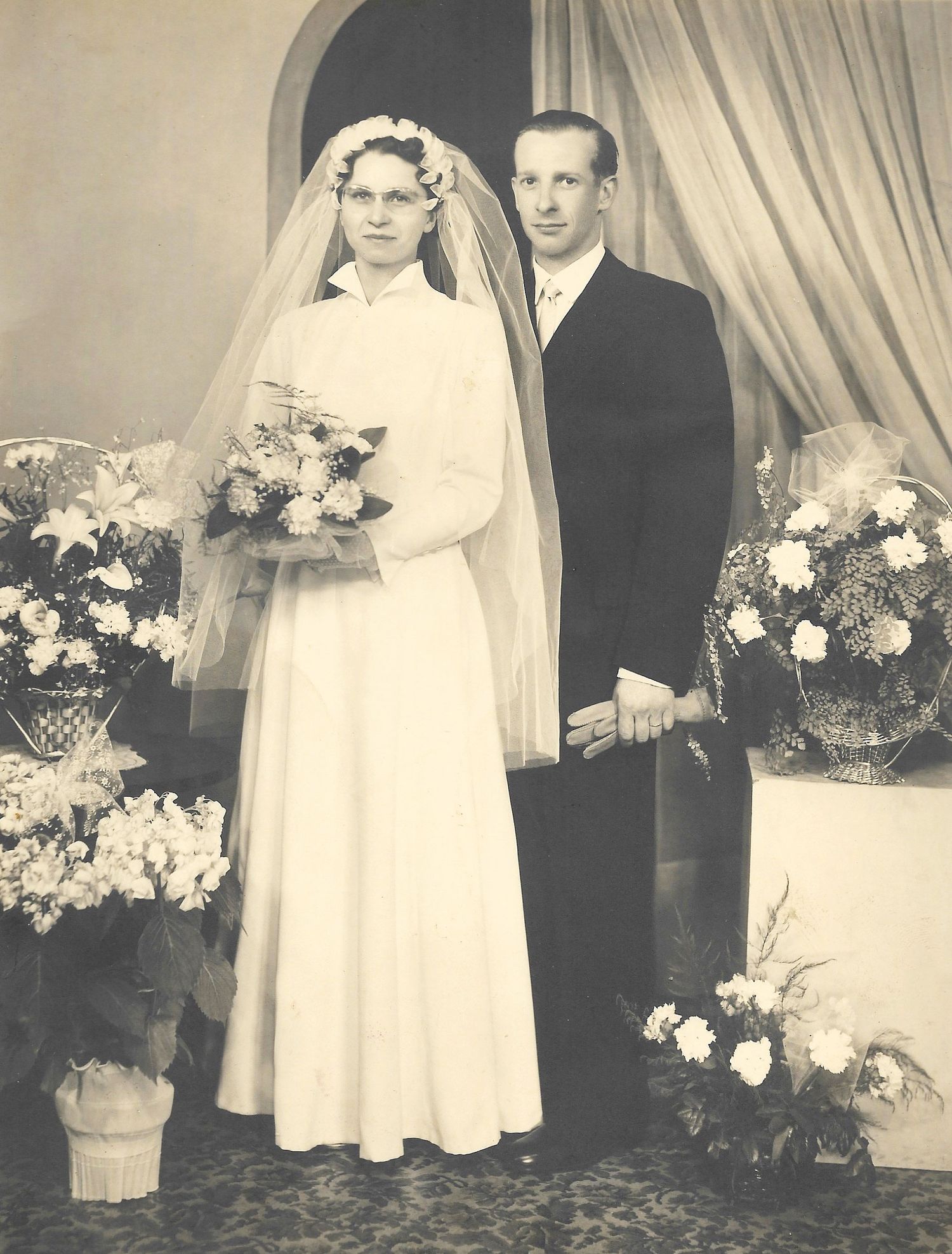 Vintage black and white wedding portrait of a bride wearing glasses and groom standing among flower arrangements.