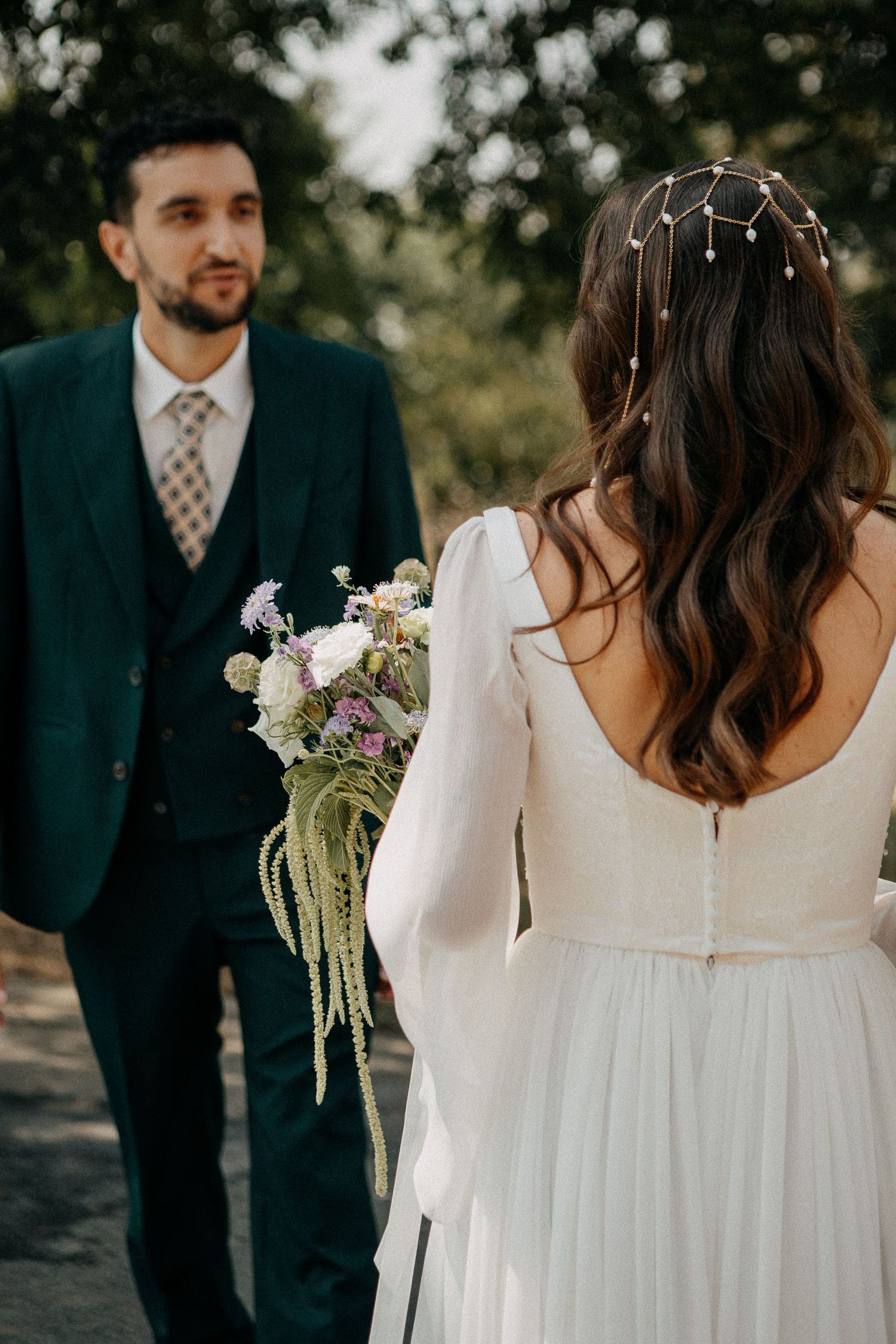 Bride in white gown with floral hair accessory faces groom in green suit holding wildflower bouquet outdoors.